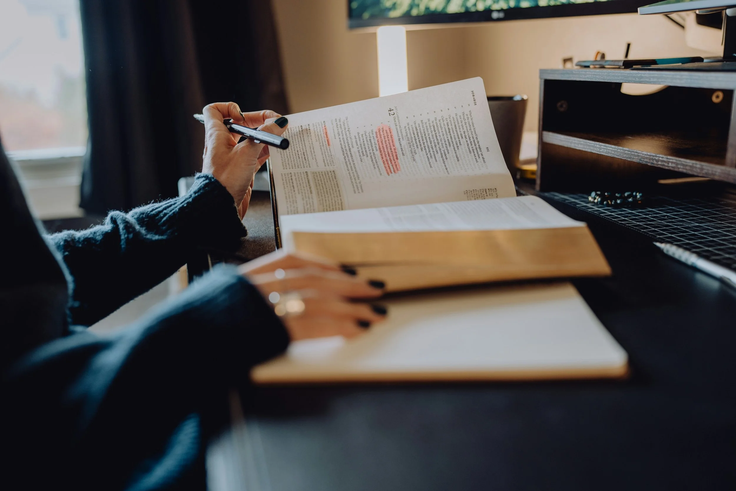 Persoon houdt een pen en een document met tekst en markeringen in een kantoorruimte, naast een computer en kantoorbenodigdheden.