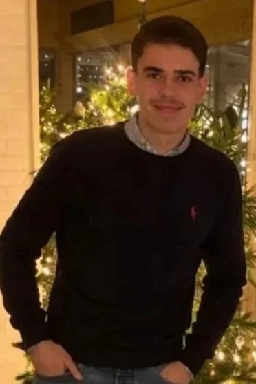 Jongeman met korte donkere haar, lichte huid en een knalroze polo logo onder een zwarte trui, staande binnen bij kerstverlichting.