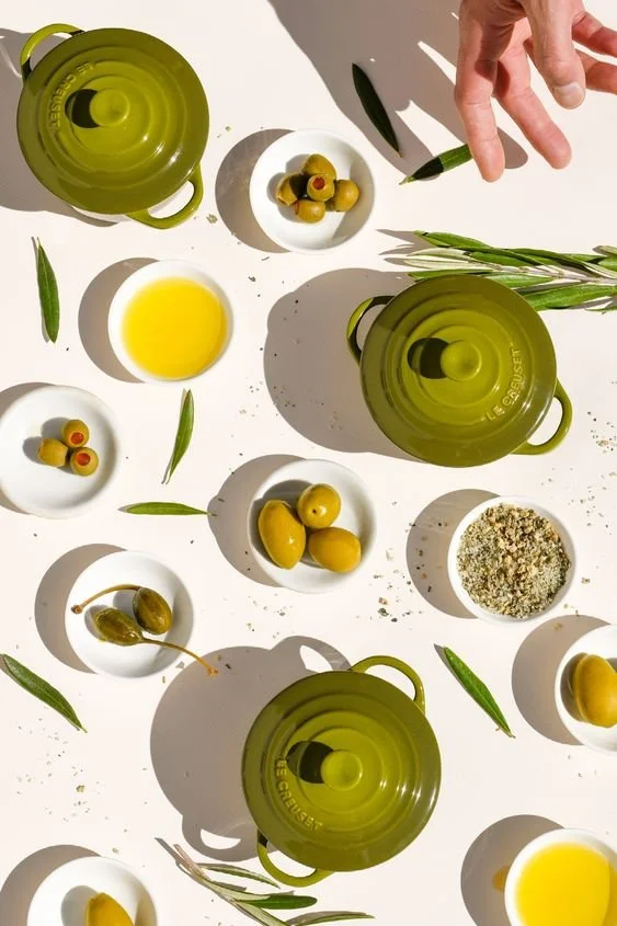 Olive oil, green olives, and various bowls with olives, seeds, salt, and herbs on a white surface, with a hand holding a green pen and green leaves scattered around.
