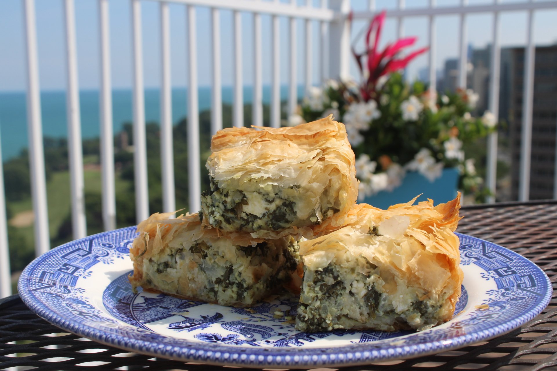 Slices of savory layered pie with phyllo dough crust on a decorative blue-and-white plate, set on an outdoor table with a balcony railing in the background and a view of distant buildings and greenery.