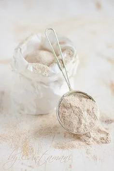 A metal scoop filled with white powder, with a container of more white powder in the background on a light-colored surface.