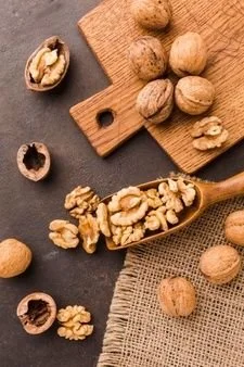 Walnuts on a cutting board and spoon, some in shells, some shelled, on a dark surface and fabric.