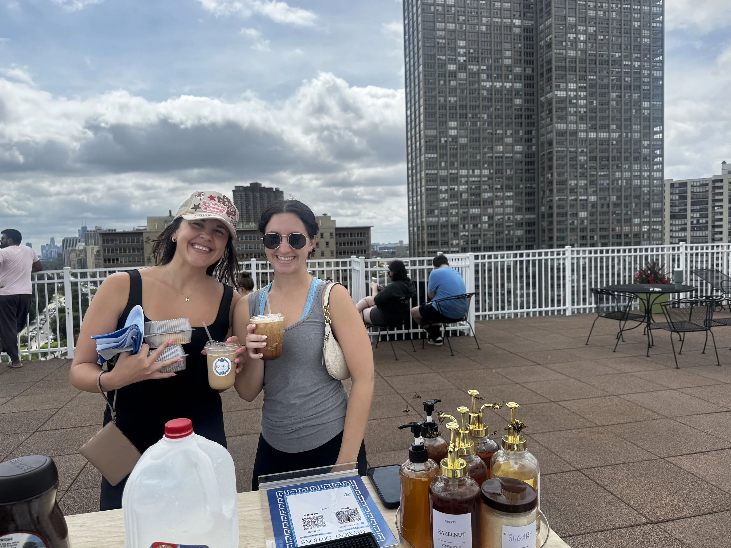 Two women smiling at a rooftop market stall with bottles of flavored syrups and a city skyline in the background.