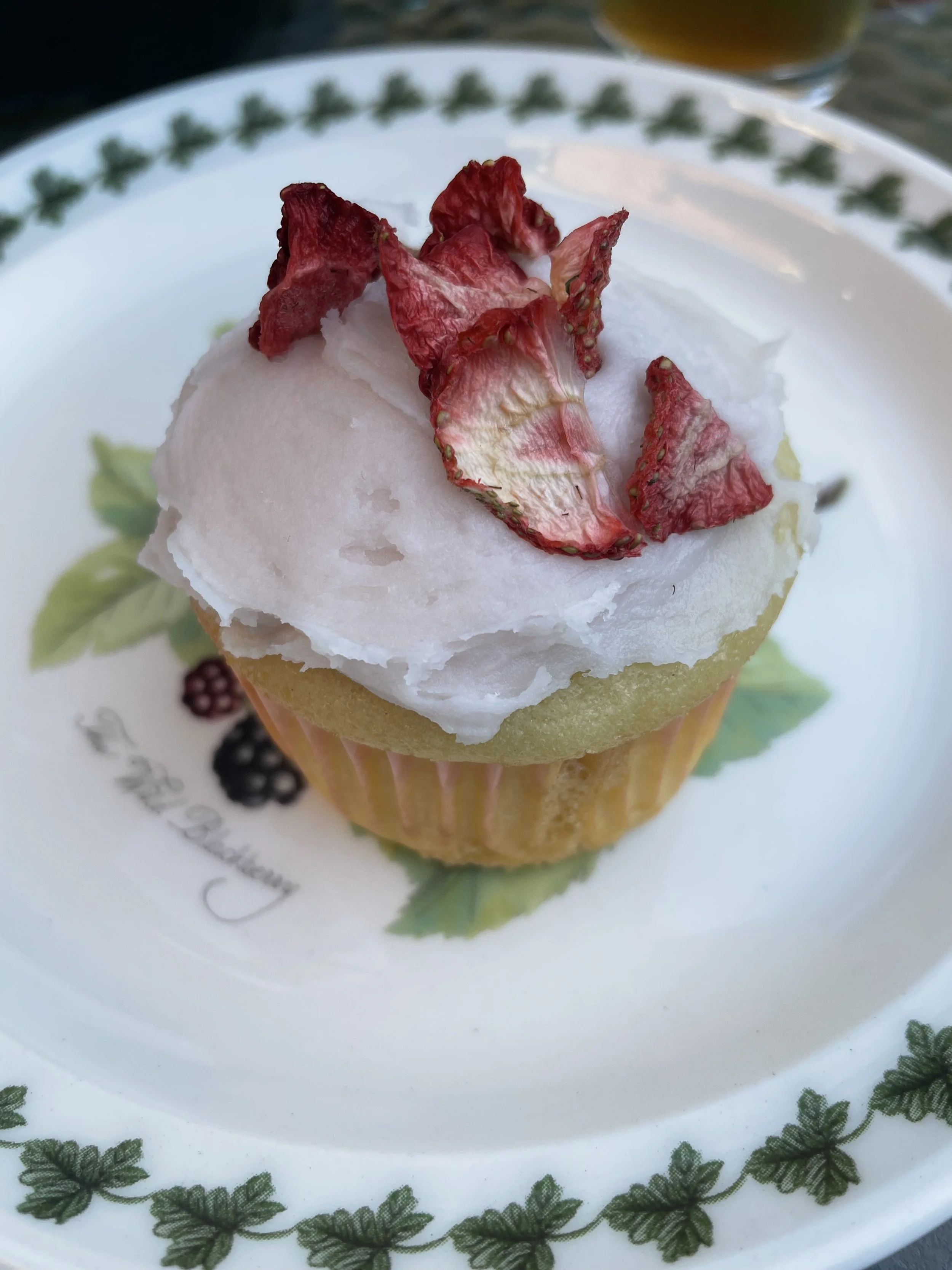 Cupcake with white frosting and dried strawberries on top, placed on a decorative plate with a green leafy border.