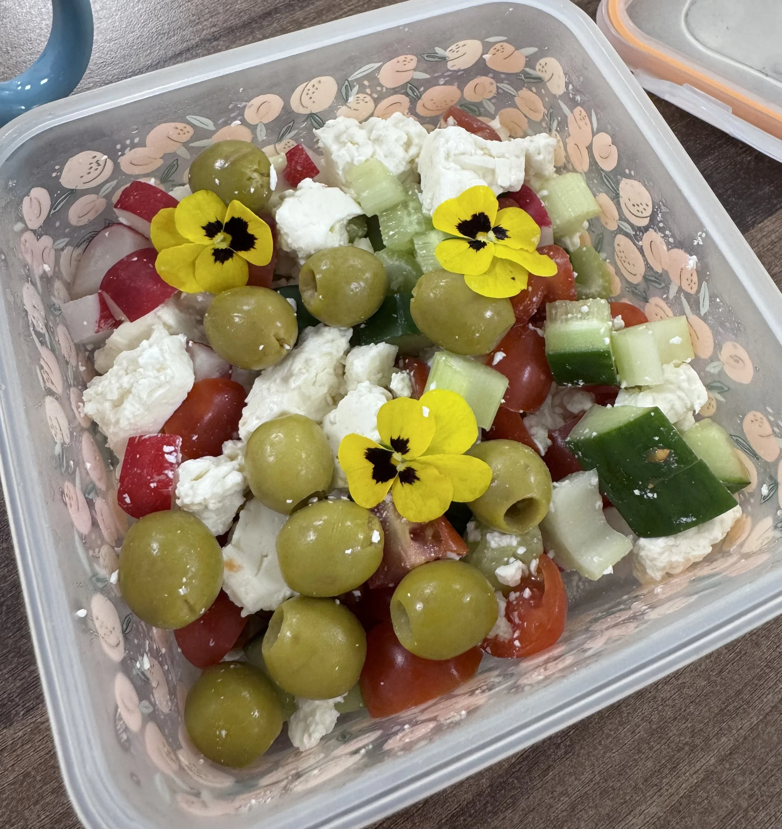 A plastic container filled with a colorful salad of green grapes, chopped tomatoes, celery, feta cheese cubes, red bell pepper, and garnished with yellow mustard flowers on top.