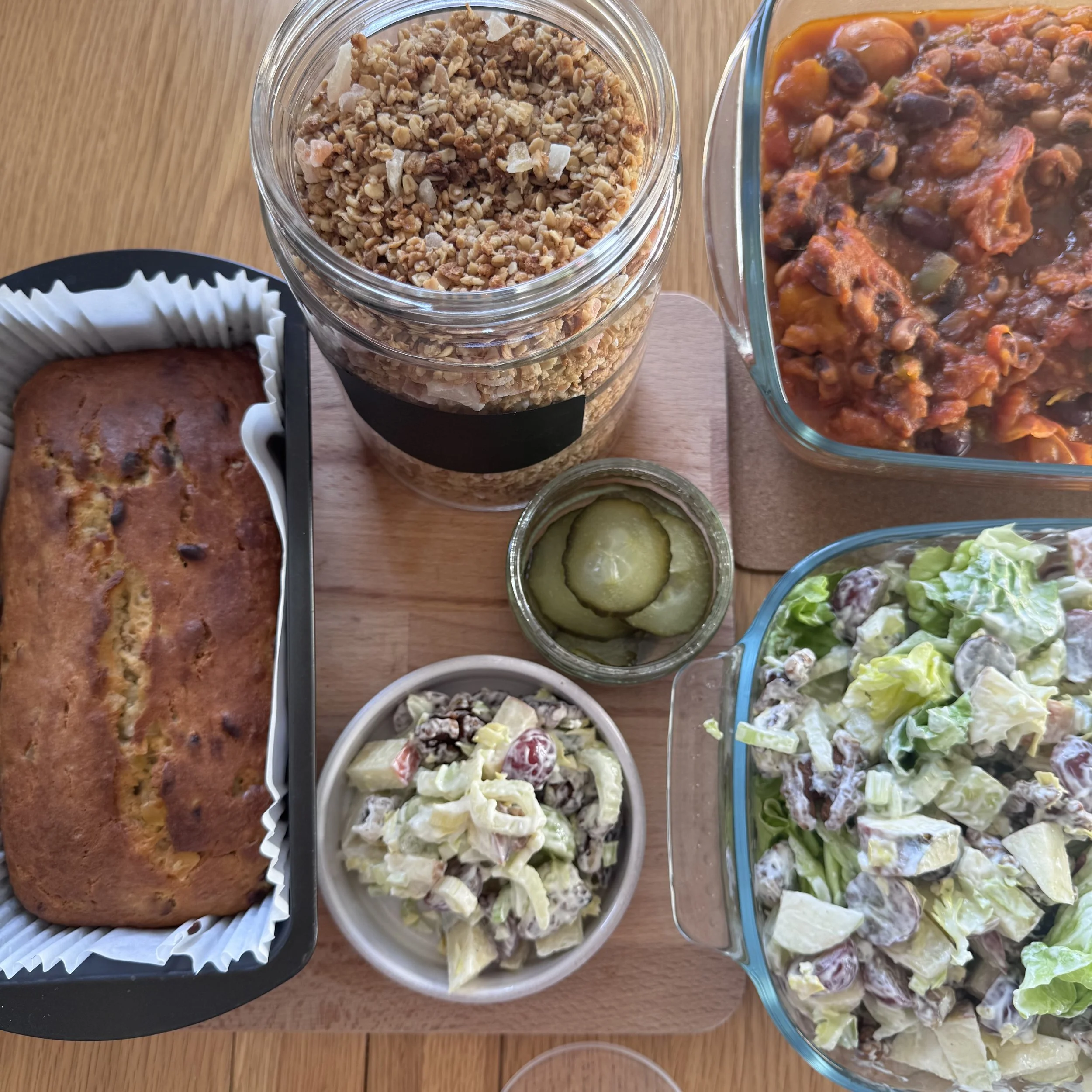 Assorted dishes including a loaf of bread, chopped nuts or granola in a jar, a vegetable or bean chili in a glass container, and bowls of salad and pickles on a wooden surface.