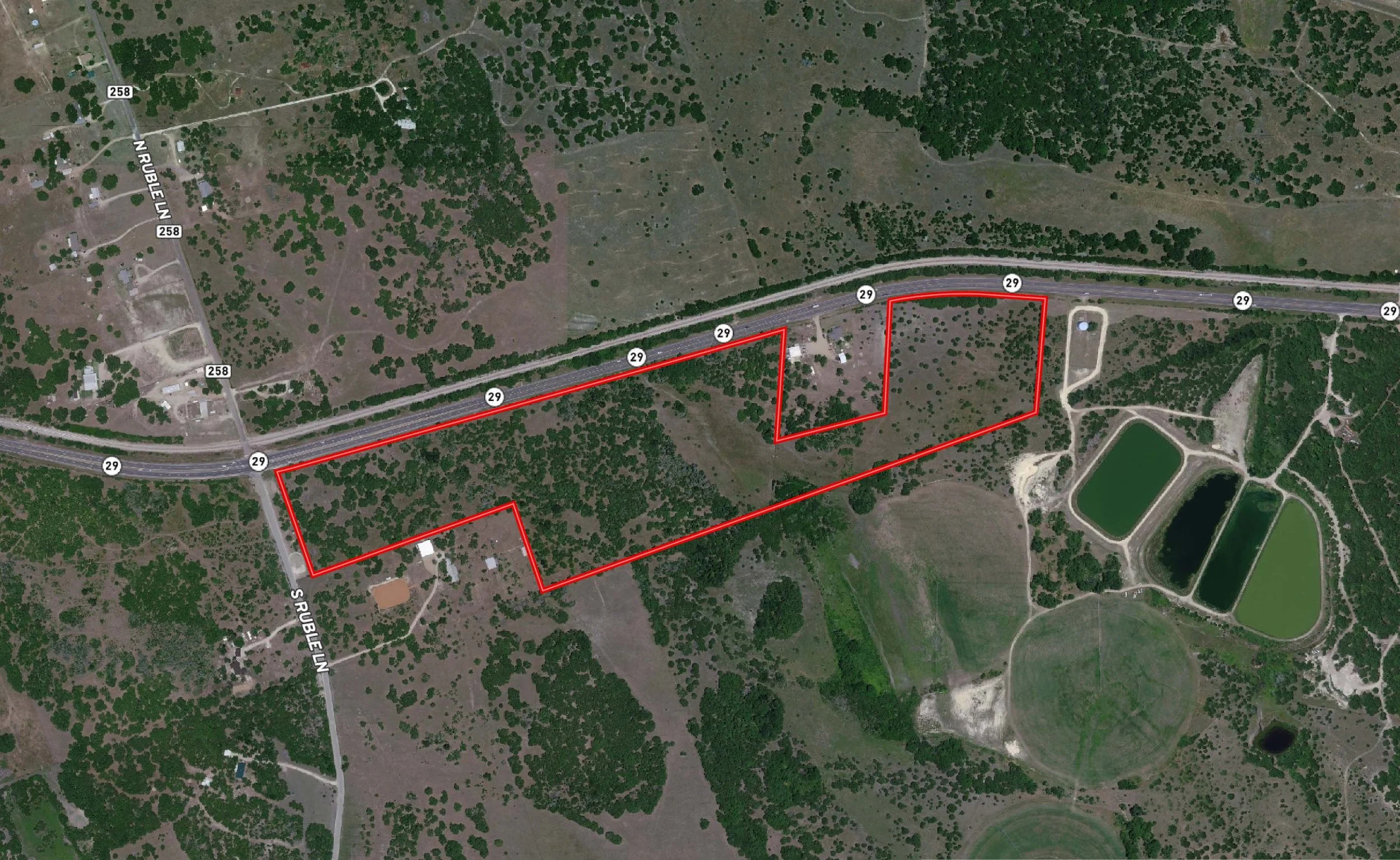 Satellite view of a rural area with a highway running horizontally across the image. Red boundary lines mark multiple plots of land surrounding the highway, scattered houses are visible, and there is a pond or water reservoir on the bottom right corner.