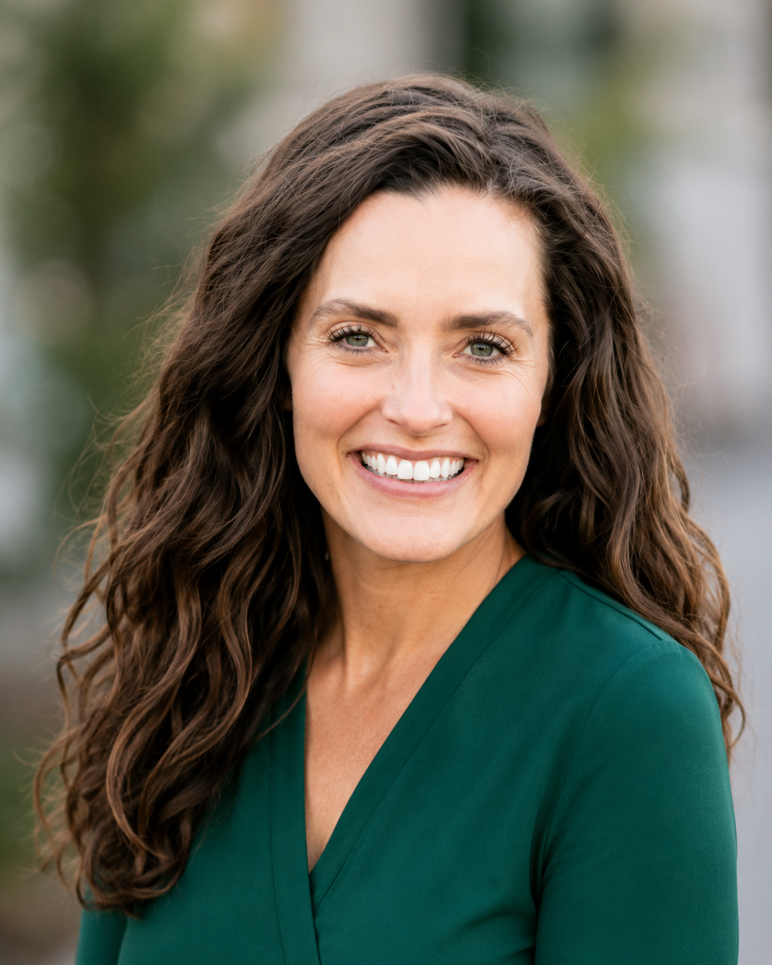 A woman with long, wavy brown hair smiling outdoors, wearing a green top.
