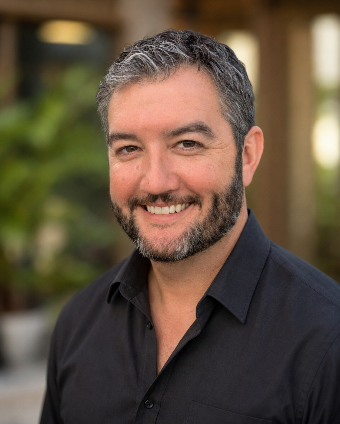 A smiling man with short, dark, and graying hair and a beard, wearing a black shirt, standing outdoors with a blurred natural background.