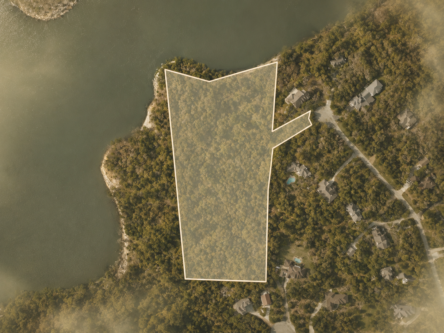 Aerial view of a residential neighborhood with a large, outlined wooded lot near a body of water.