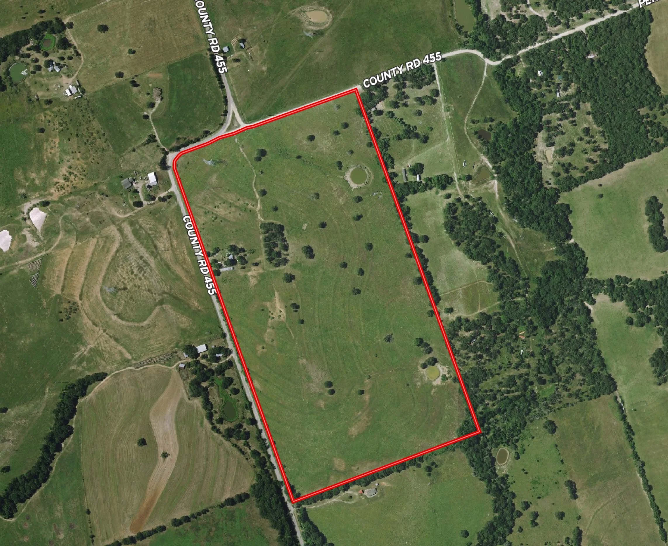 An aerial view of a rural area with open green fields, scattered trees, small ponds, and houses. Red boundary lines outline a specific parcel of land. Roads labeled 'COUNTY RD 455' and 'COUNTY RD 445' run through or near the area.