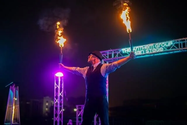 A fire performer in a vest and hat spins flaming torches on an outdoor stage at night.
