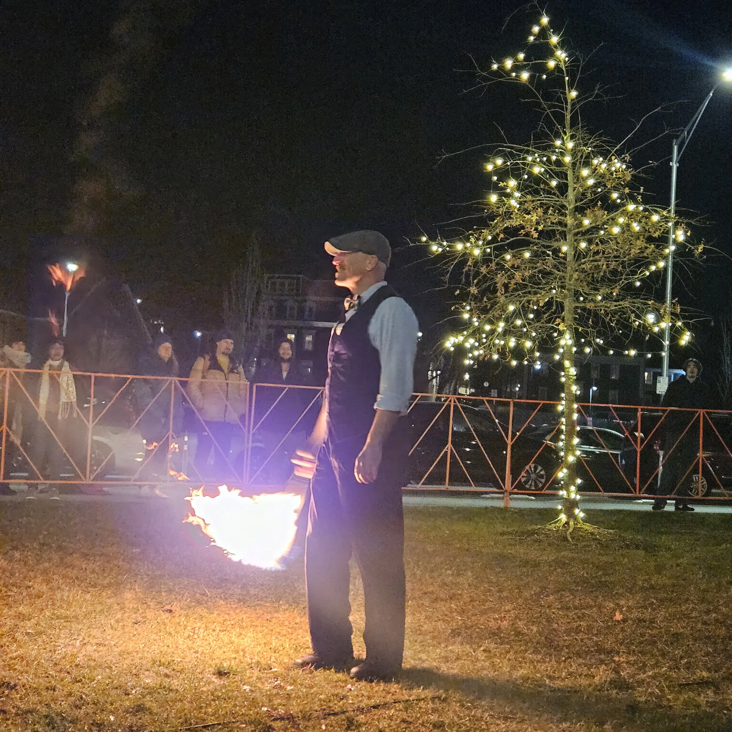 A man standing outdoors at night near a lit Christmas tree, with a lit fire pit on the ground and people in the background.