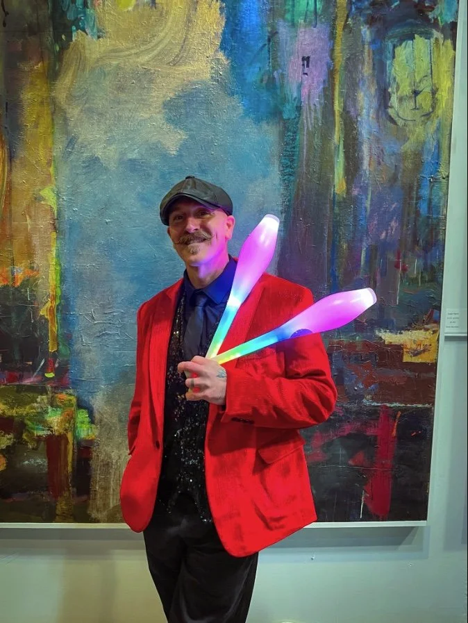Jay Moran in a red blazer, black shirt, and flat cap holding colorful LED clubs, standing in front of an abstract painting.