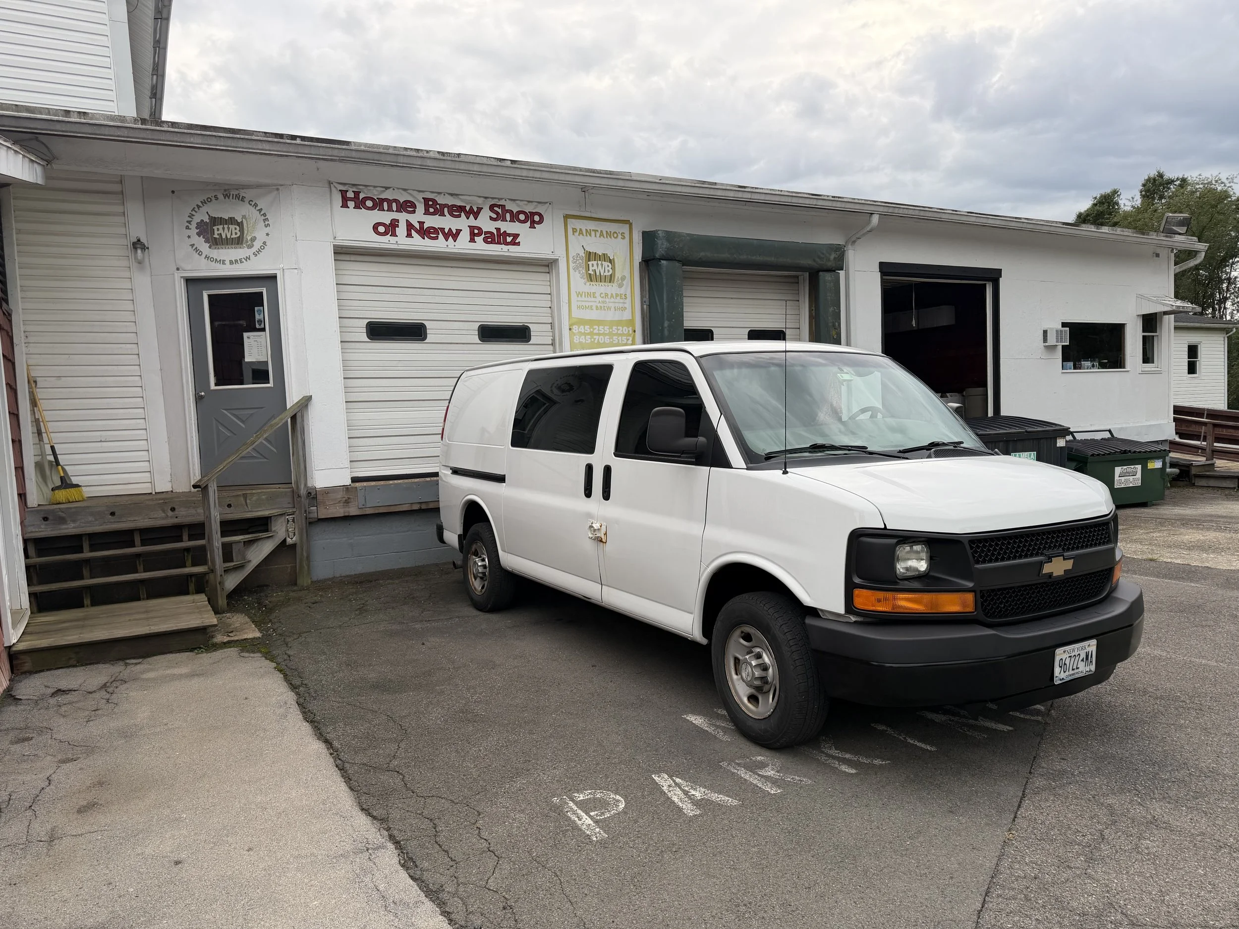 White van parked in front of a small building with a sign that reads 'Home Brew Shop of New Paltz' and advertising for Pantano's wine grapes and home brew shop. The sky is cloudy.