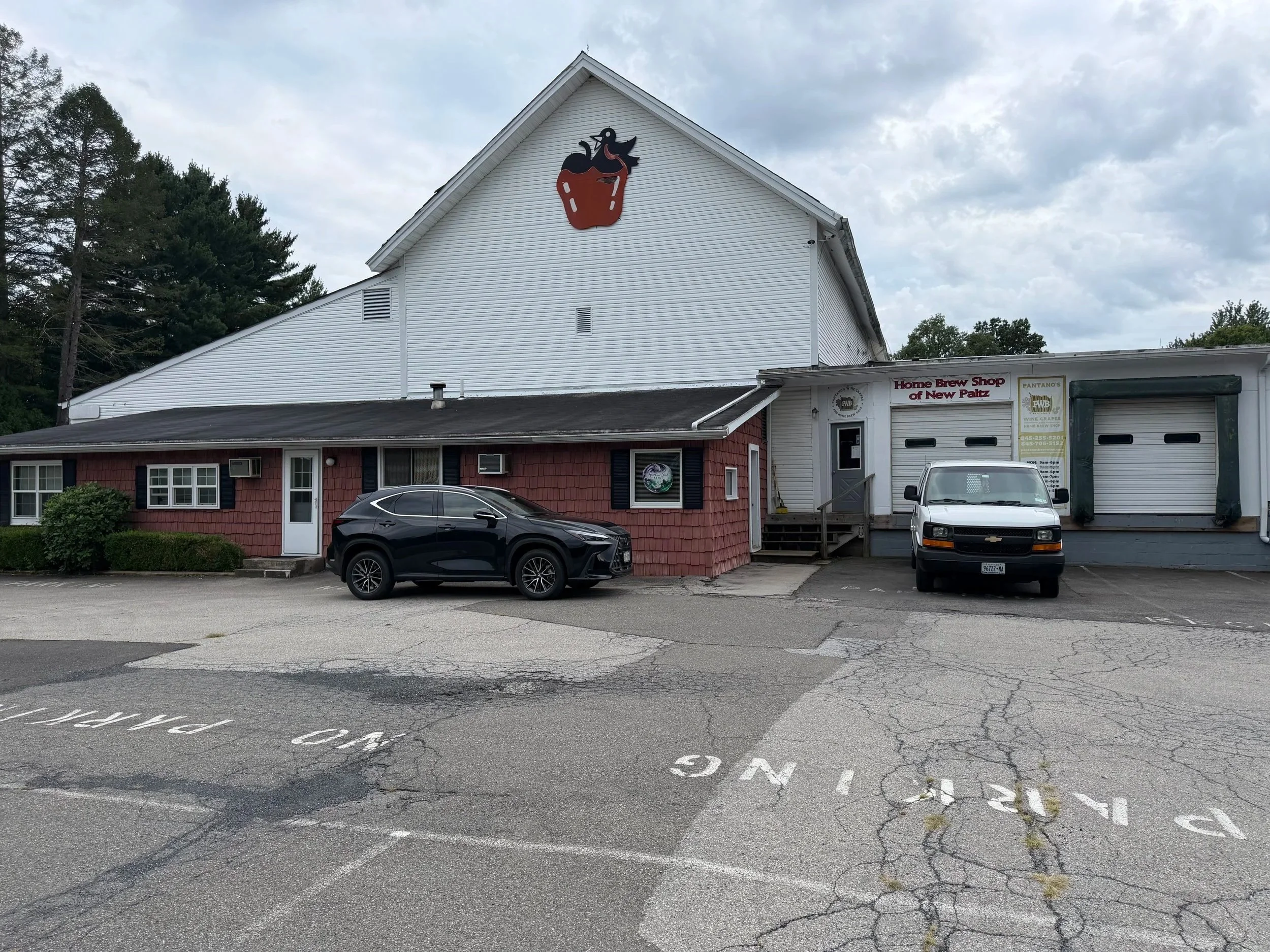 Parking lot with two cars, one black SUV and one white truck, in front of a white building with a large cupcake and ghost sign and a smaller sign for the Home Brew Shop of New Paltz.