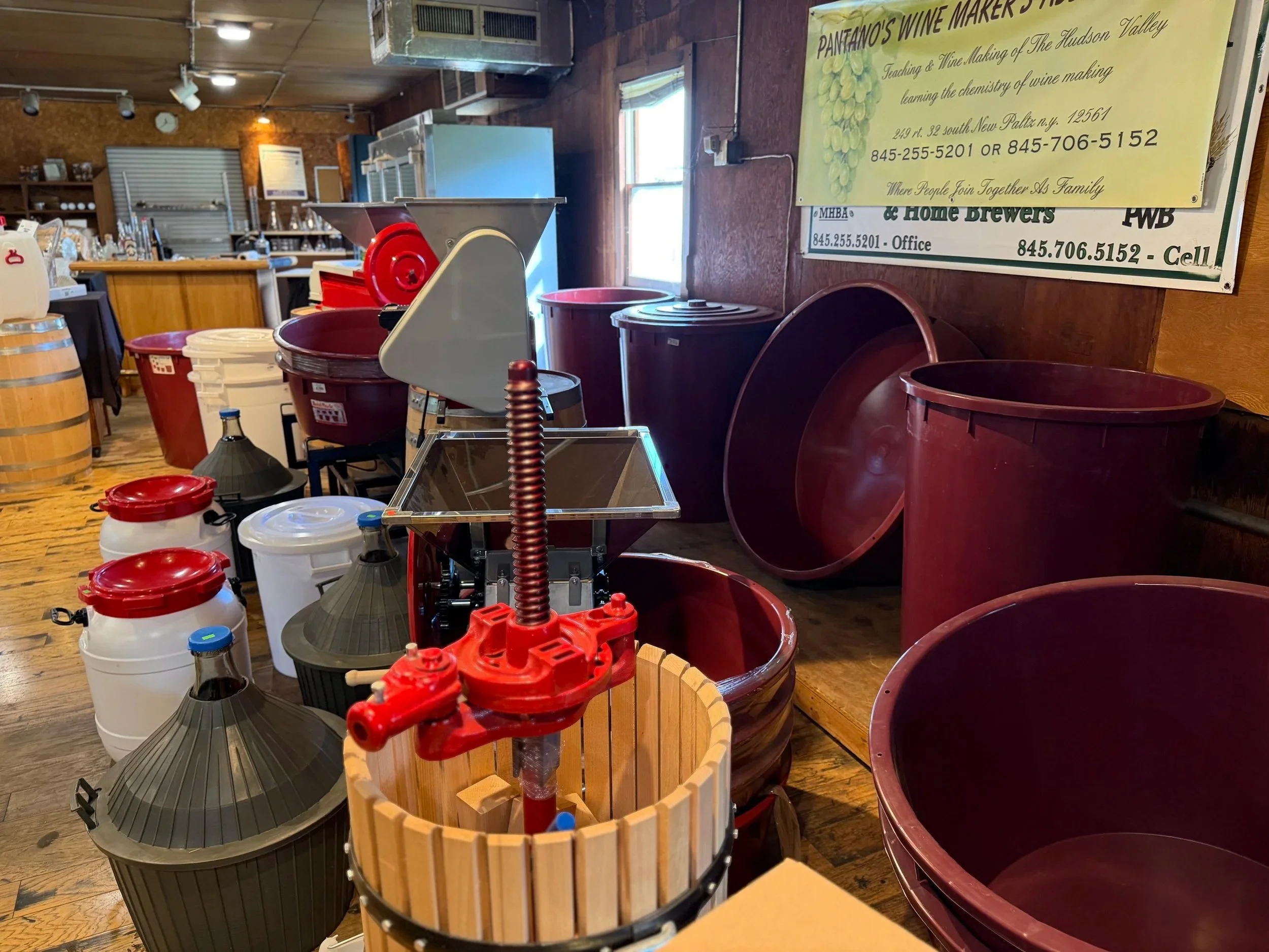 Inside a winery or wine making facility with various equipment, barrels, and containers, alongside posters on the wall related to wine making and classes.