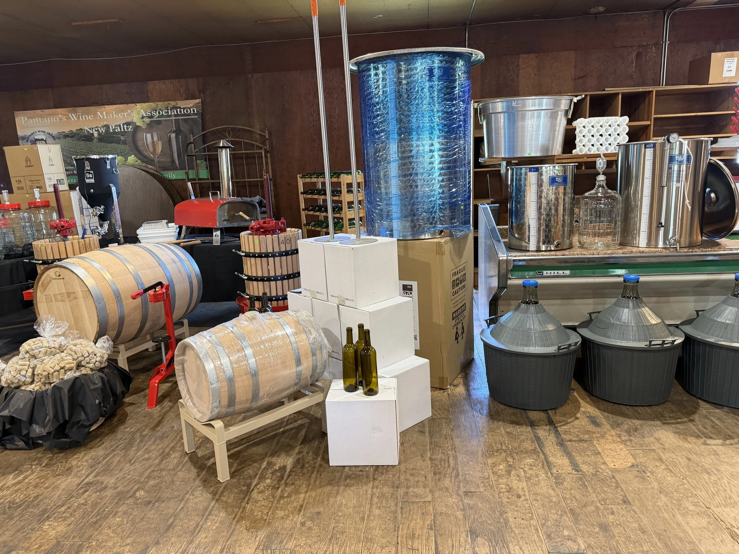 Display of wine-making equipment including a large blue tank, glass demijohns, wine barrels, and various accessories on shelves and tables at a wine studio or cellar.