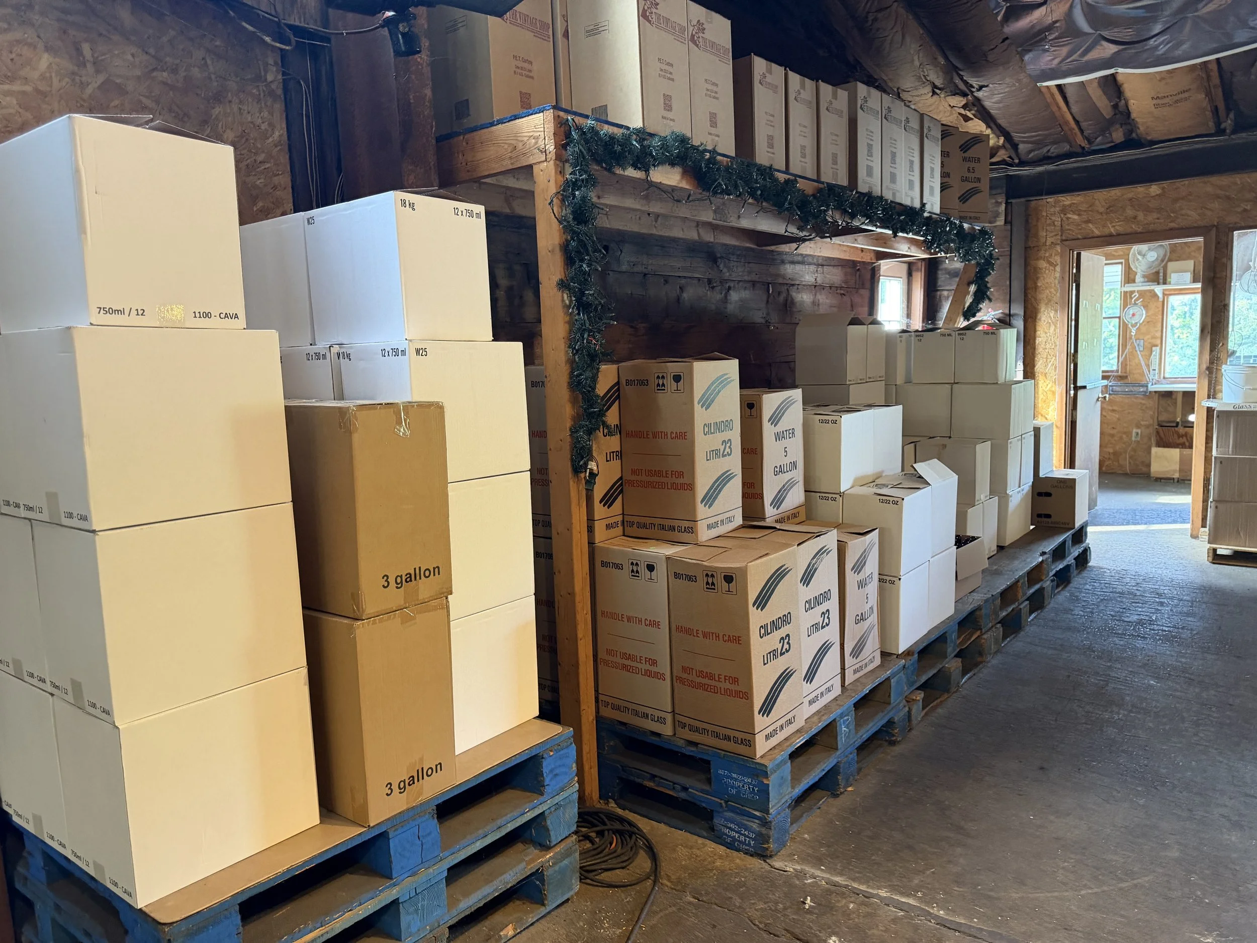 Stacked boxes of beverages including water and canned drinks in a storage room with wood walls, a window, and black ductwork on the ceiling.