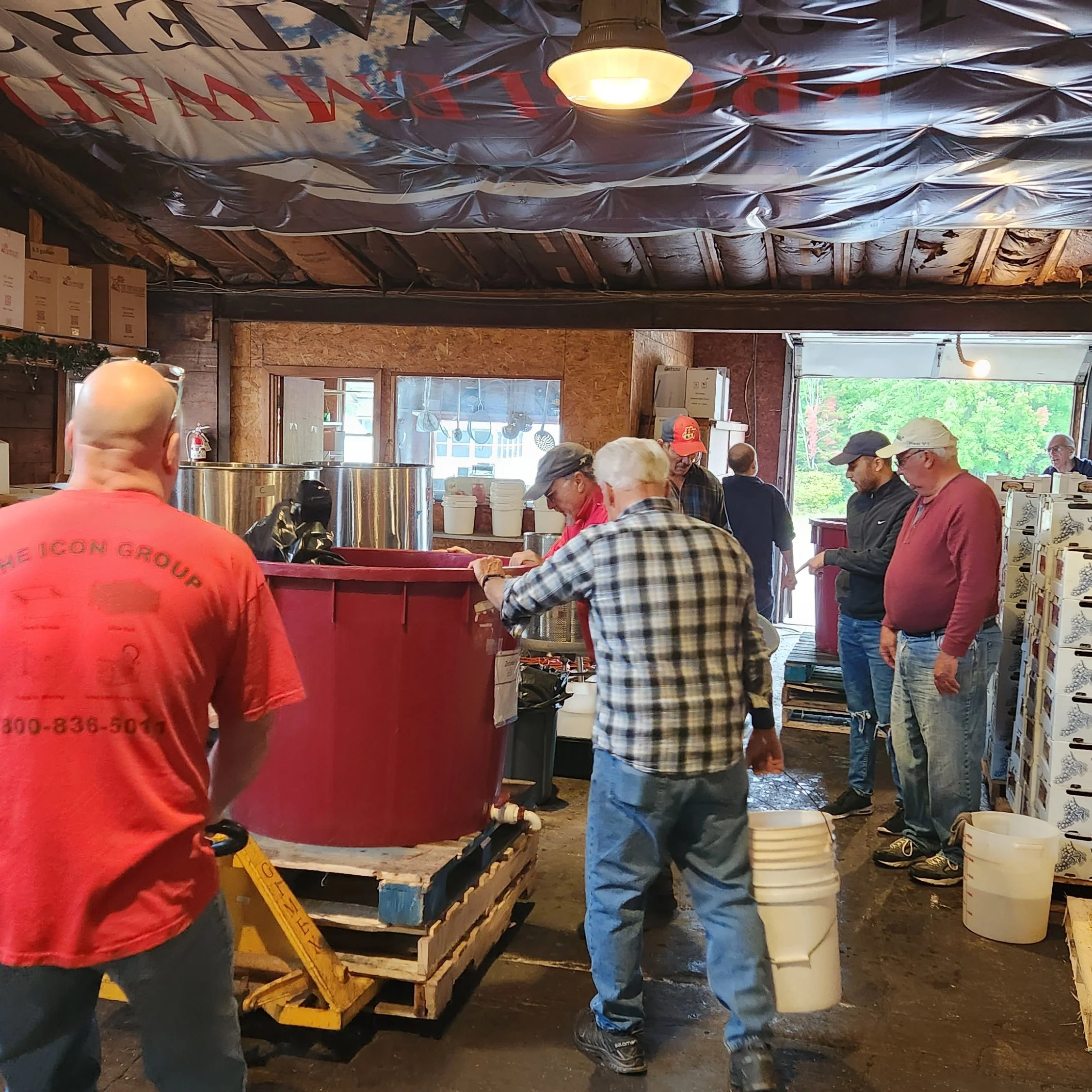 People standing in line at what appears to be a market or food stall, with cardboard boxes, buckets, and wooden pallets around. Some individuals are wearing hats and aprons, indicating they might be vendors or customers.