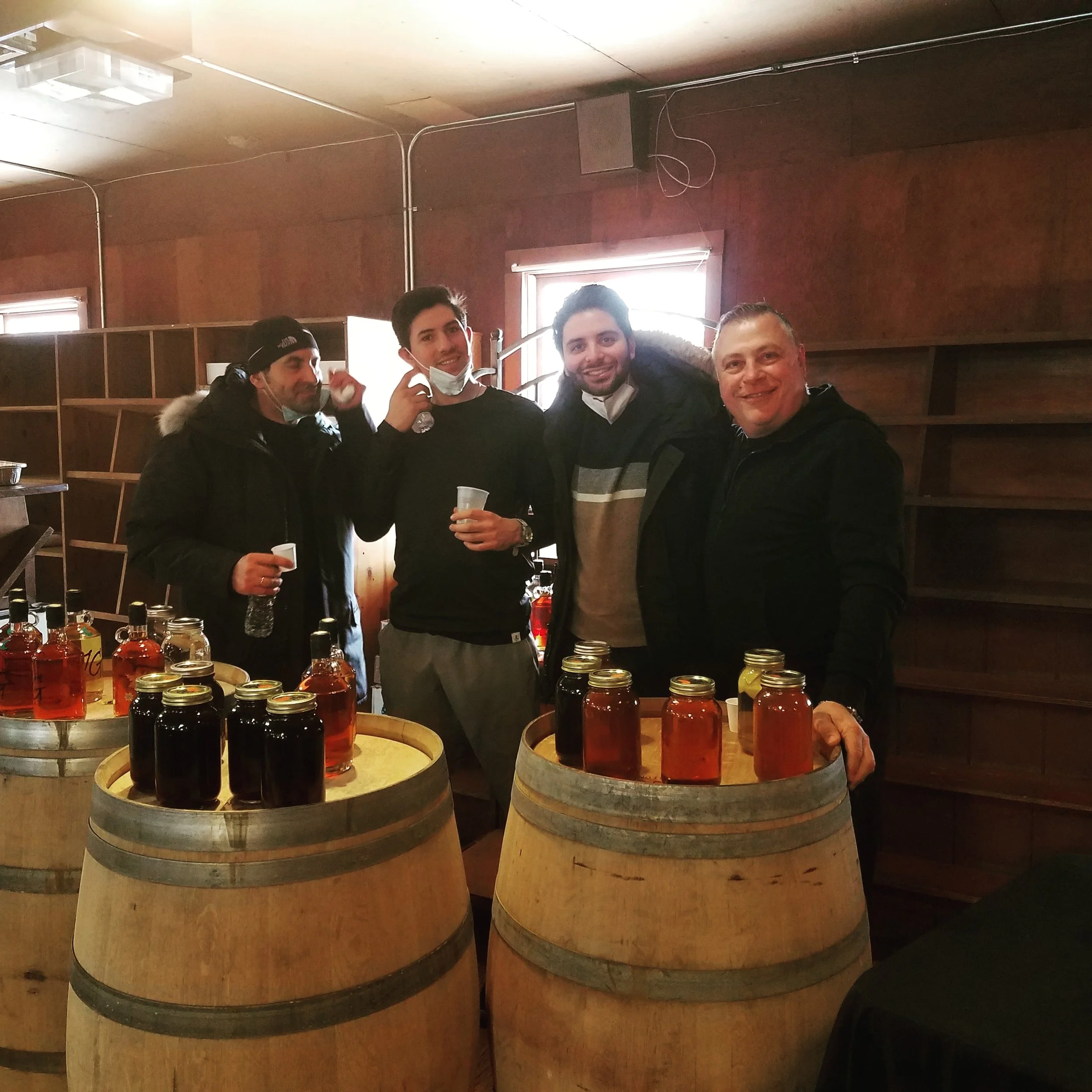 Four men standing behind a table with jars of liquids inside wine barrels, inside a rustic wooden room with two small windows and a ceiling with exposed pipes.