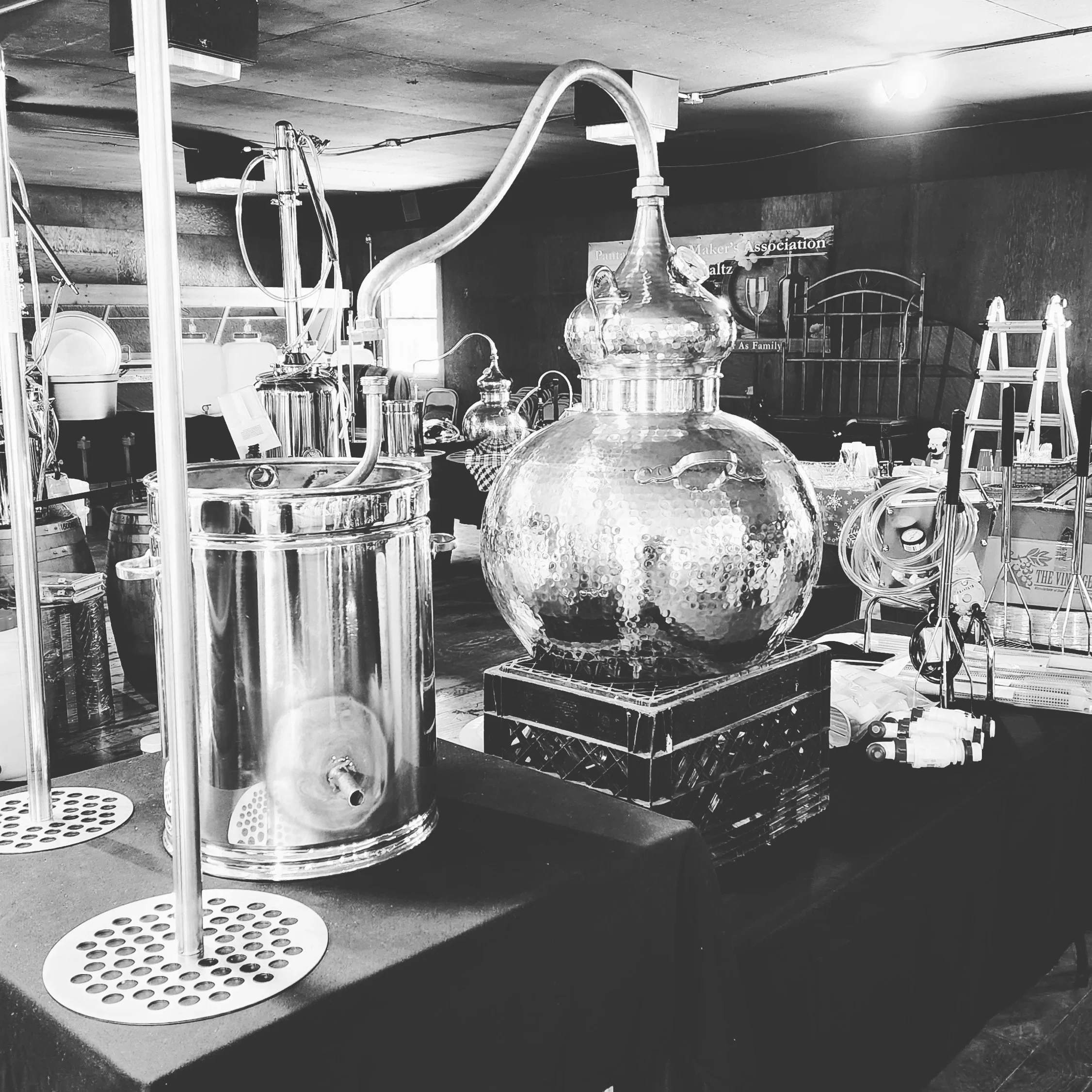 Black and white photo of brewing equipment including large glass distillation apparatus, metal tanks, hoses, and related tools in a workshop or brewery setting.