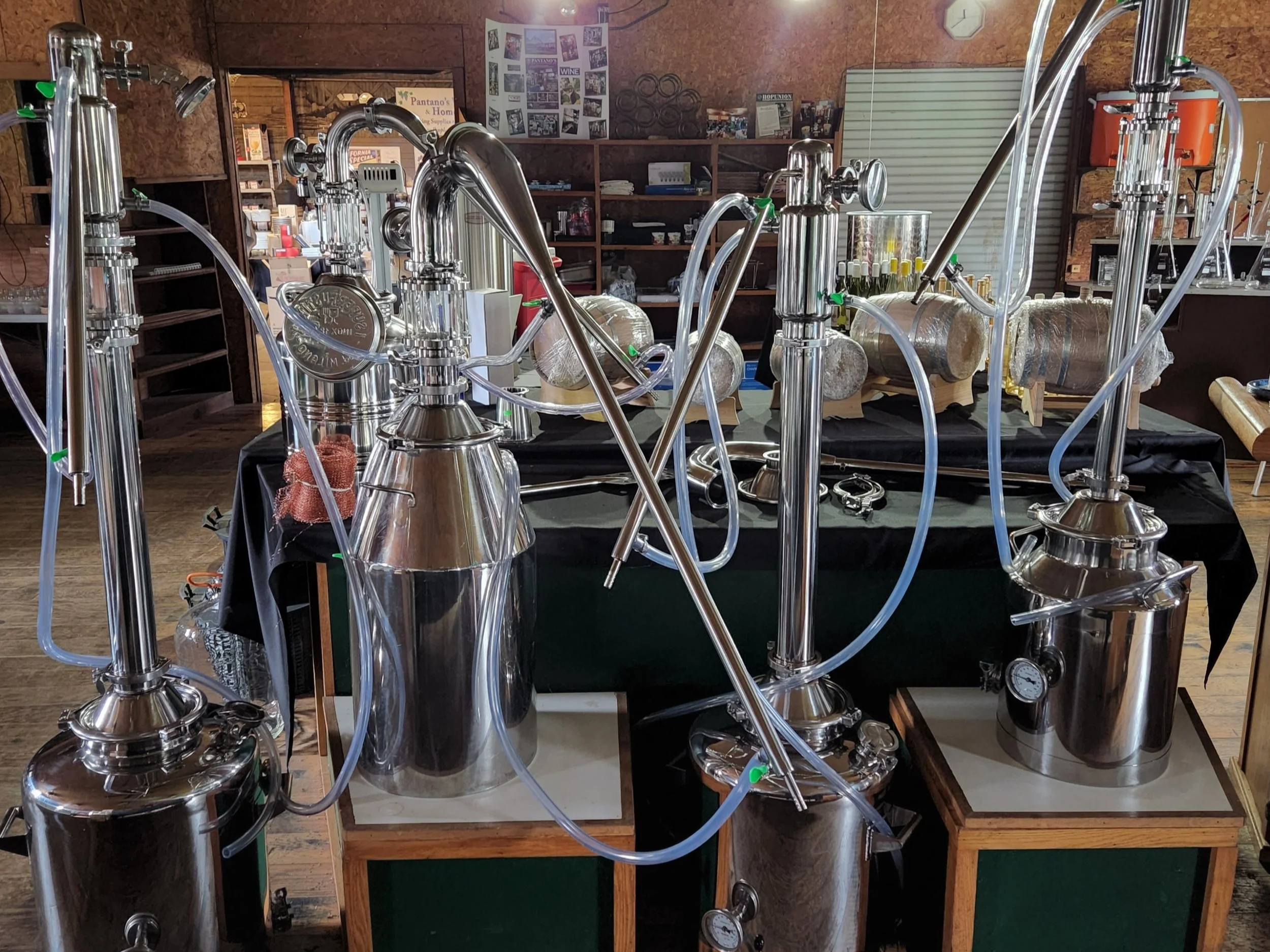 An indoor display of stainless steel distillation apparatus including multiple connected columns, tubes, and containers with a background of wooden shelves filled with various items and posters on the wall.