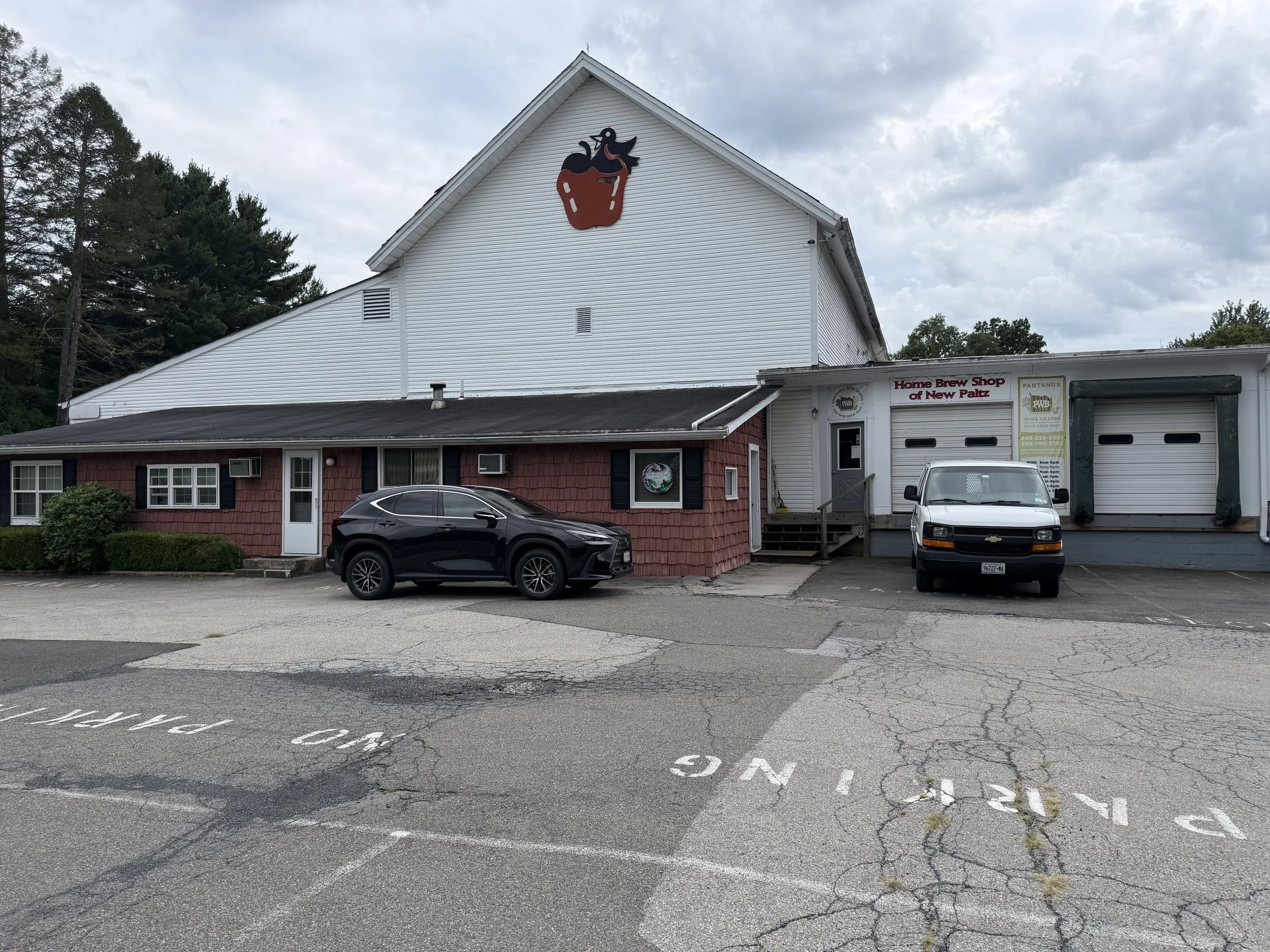A white building with a cup cake logo on the upper part, and a sign indicating it is a Home Brew Shop. There are parked cars in front of the building and some trees in the background under a cloudy sky.