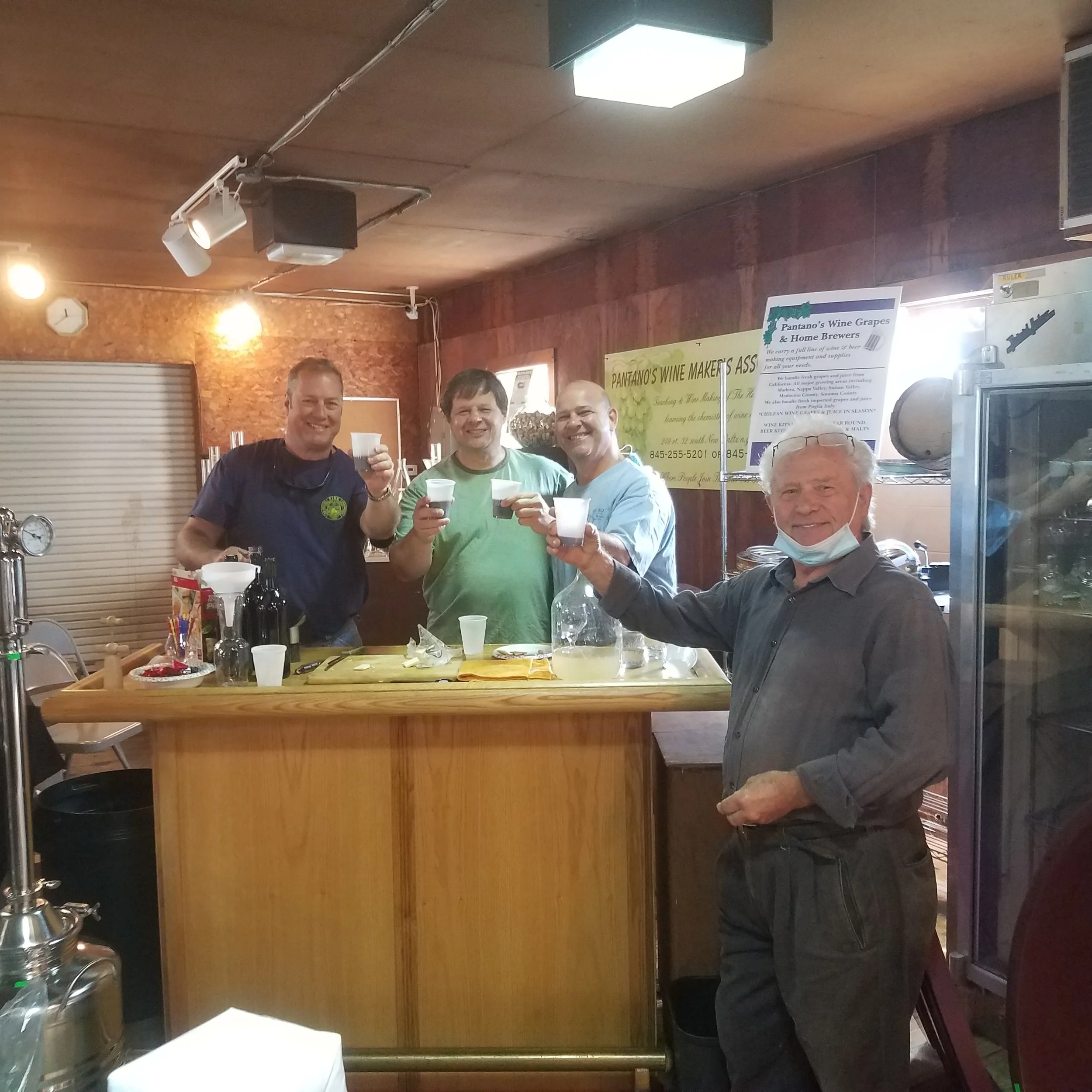 Three men standing behind a bar counter, smiling and holding cups in a cozy indoor setting with wooden walls, bright lights, and various signs and bottles in the background.