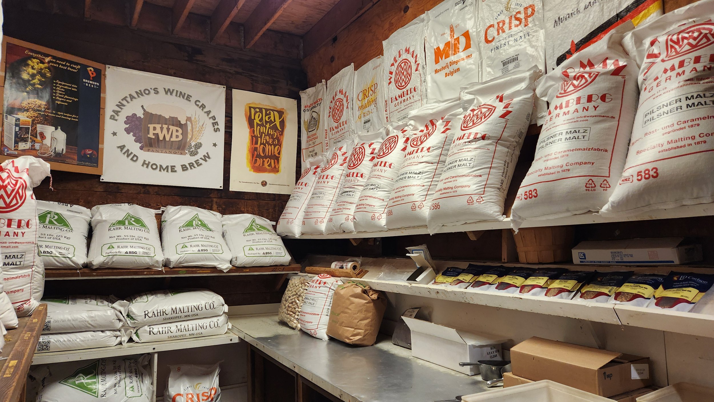 Shelves filled with bags of malt, brewing ingredients, and posters related to home brewing in a storage room.