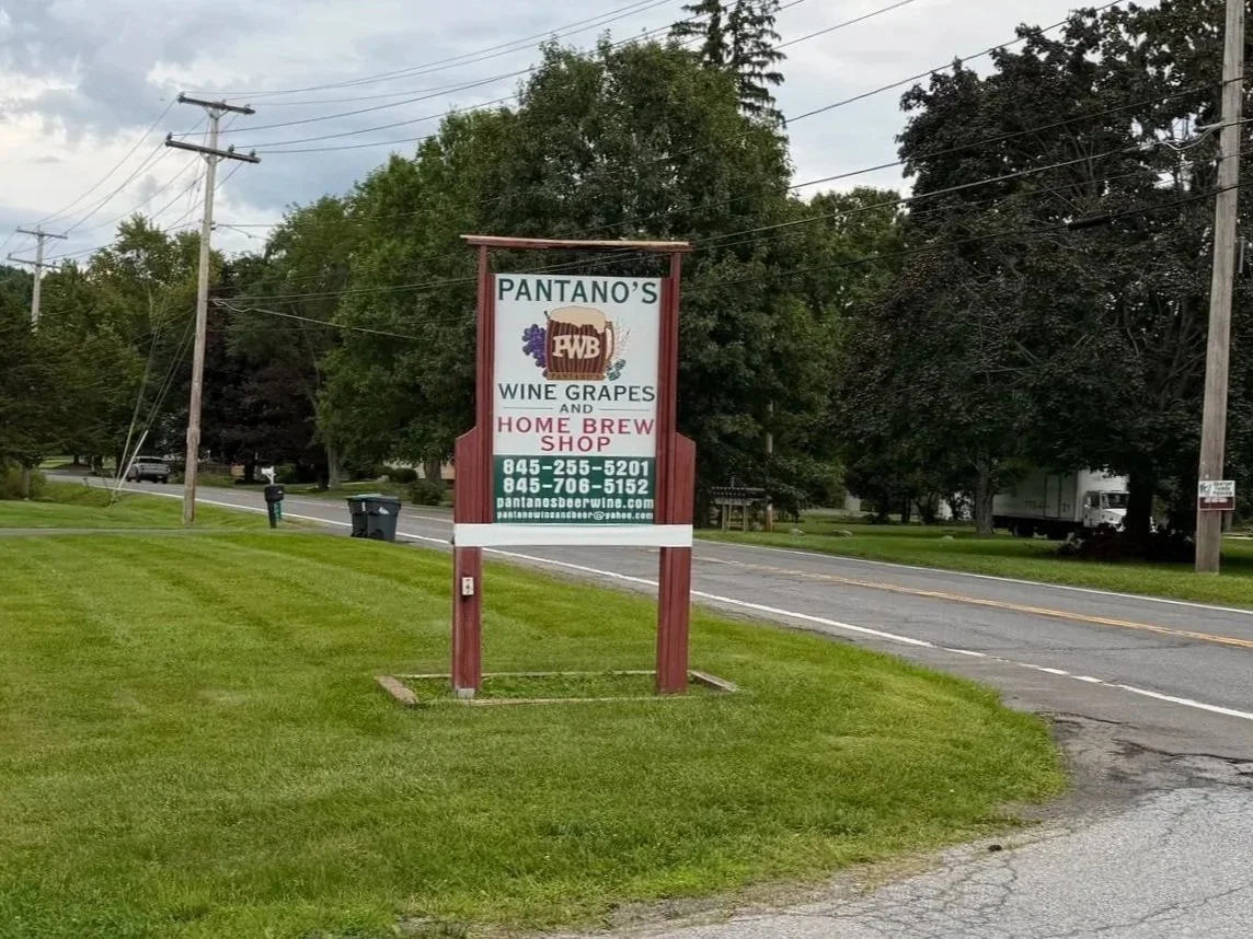 Sign for Pantano's wine grapes and home brew shop with contact information, on a grassy roadside near a road with trees and utility poles in the background.