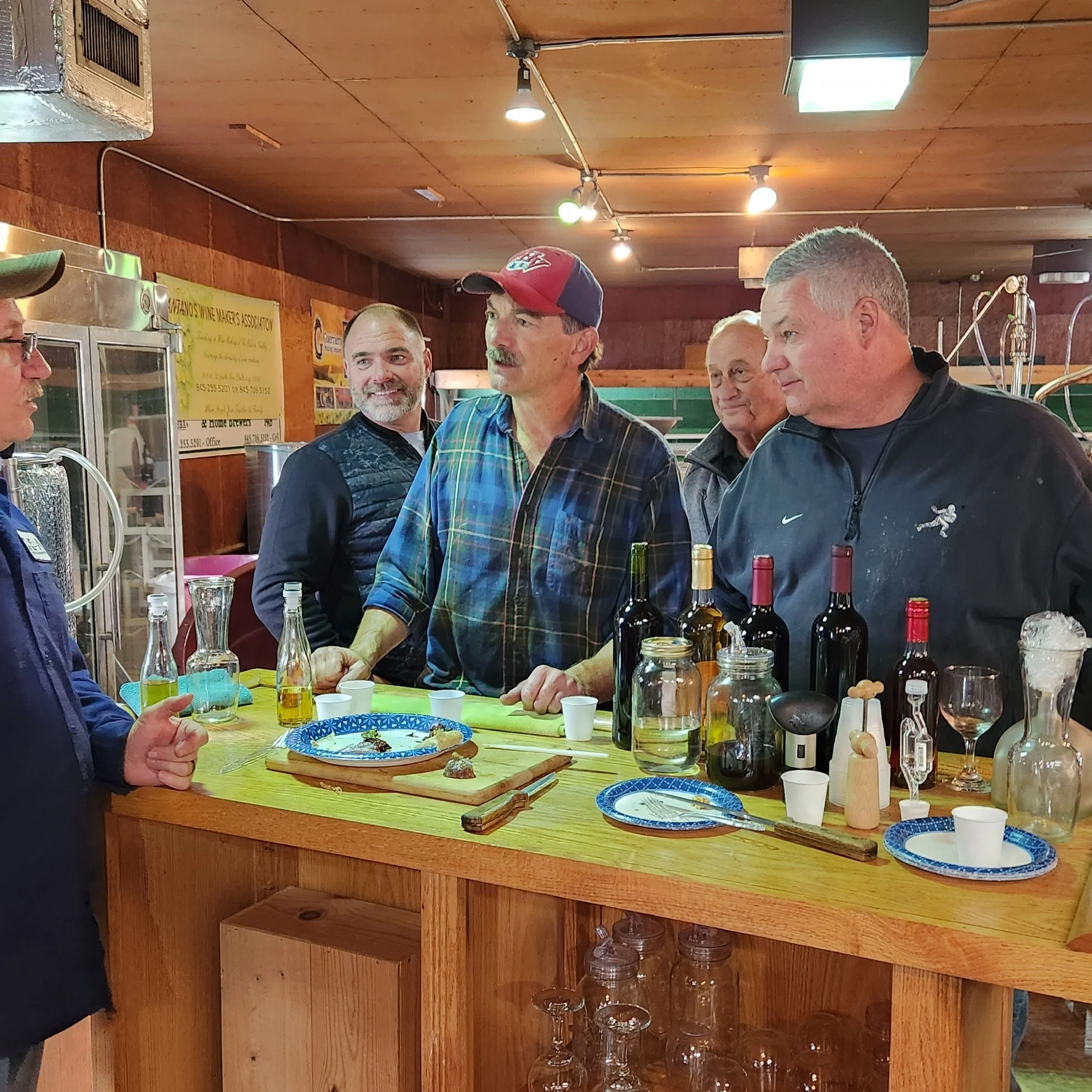 A group of men gathered around a wooden bar or counter with wine bottles, glasses, and appetizers, in a room with wood-paneled ceiling and walls, warm lighting, and some posters on the wall.