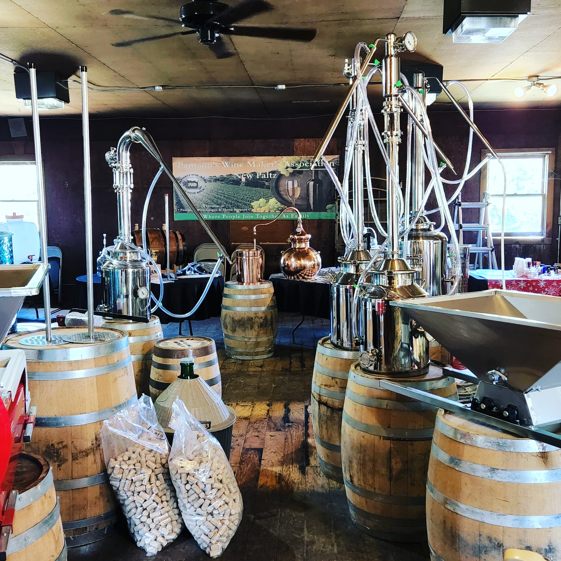 Interior of a winery with several large wooden barrels used for aging wine, a brewing or distilling apparatus with shiny metallic pipes and containers, and a poster in the background advertising a wine association in New Paltz.