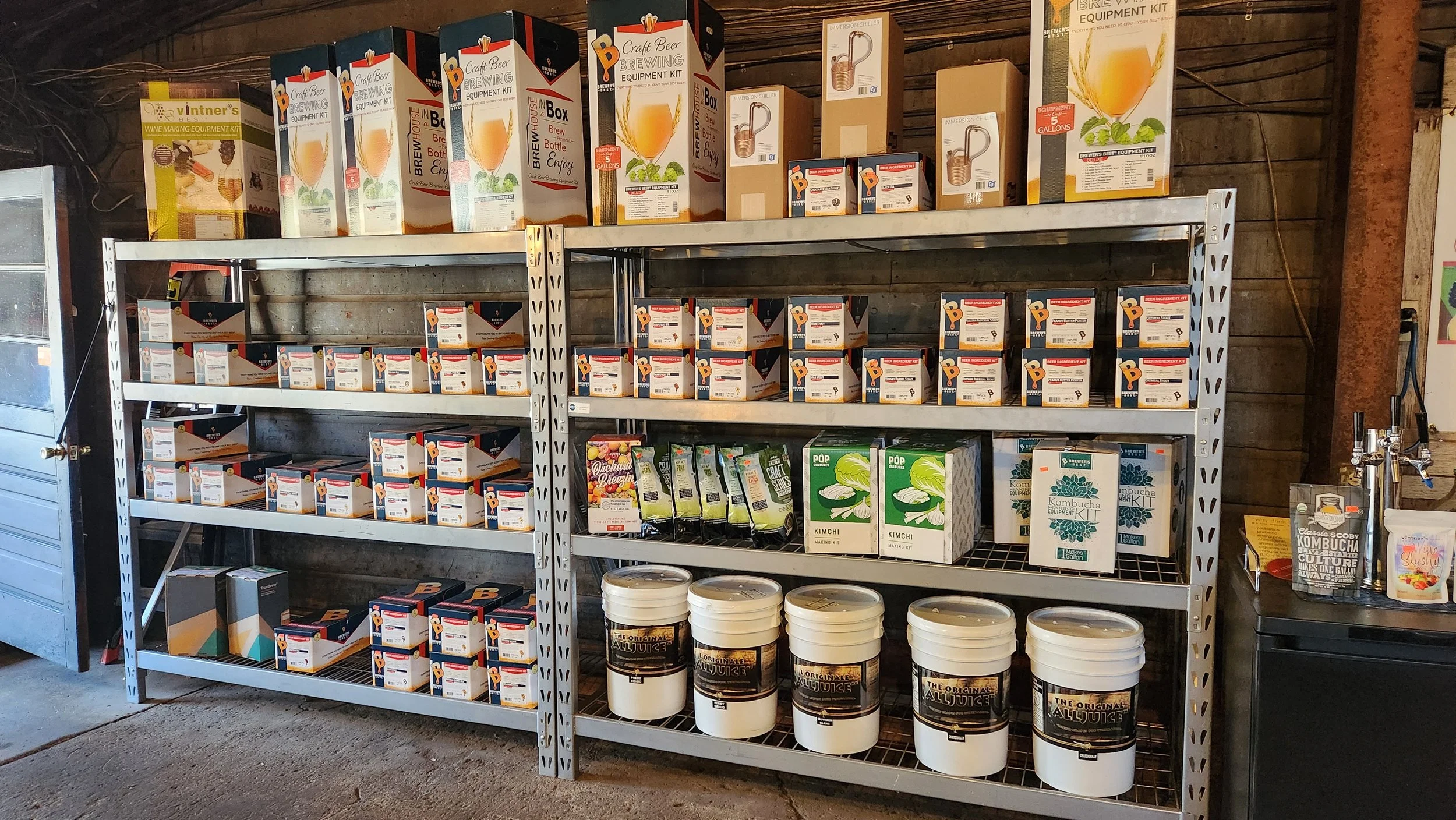 Steel shelving unit stacked with beer brewing, wine making, kimchi, kombucha, and other fermentation kit boxes in a rustic setting.