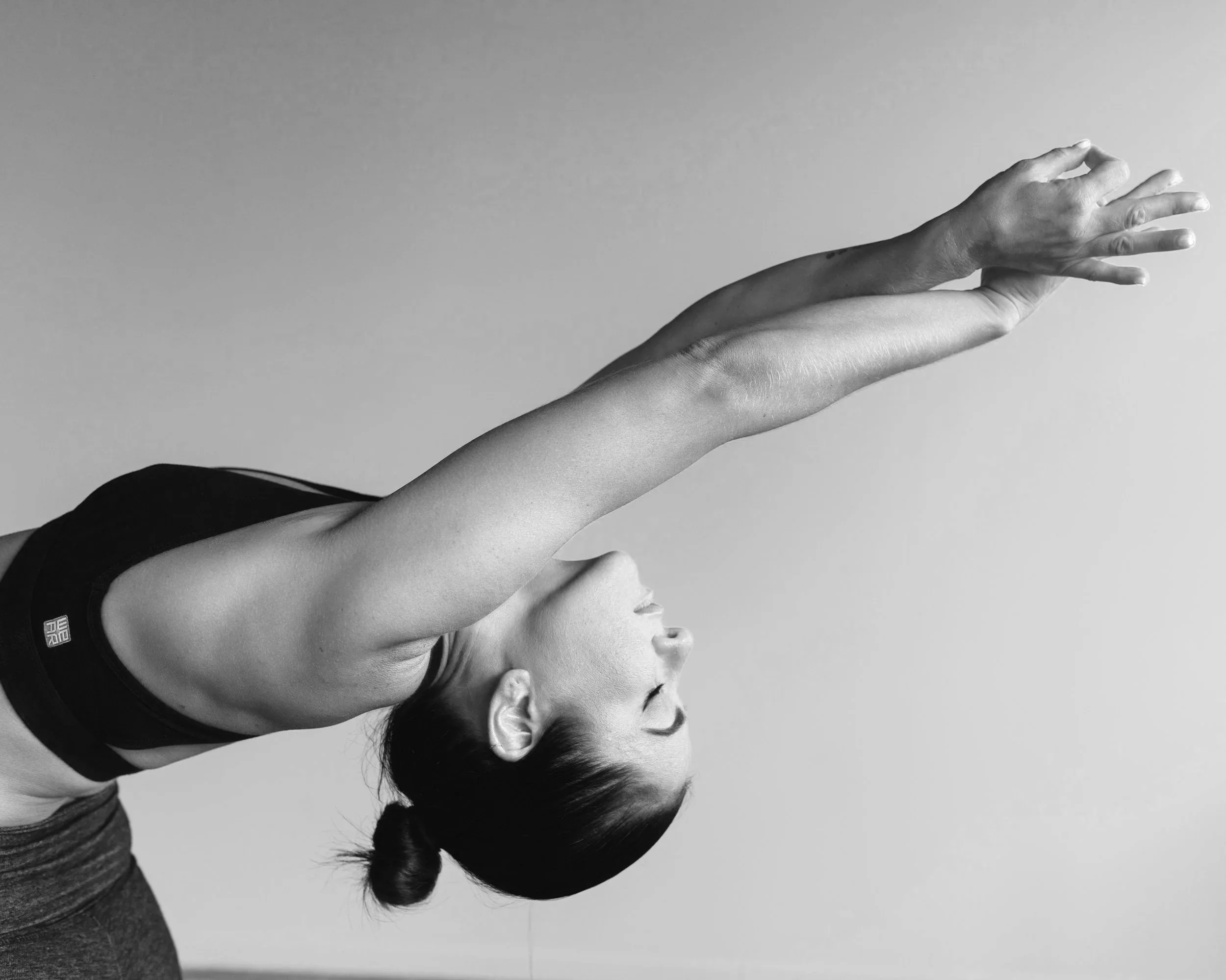 Une femme en position de yoga ou d'étirement, vue de profil, avec les bras tendus vers l'avant et la tête inclinée en arrière. La photo est en noir et blanc.