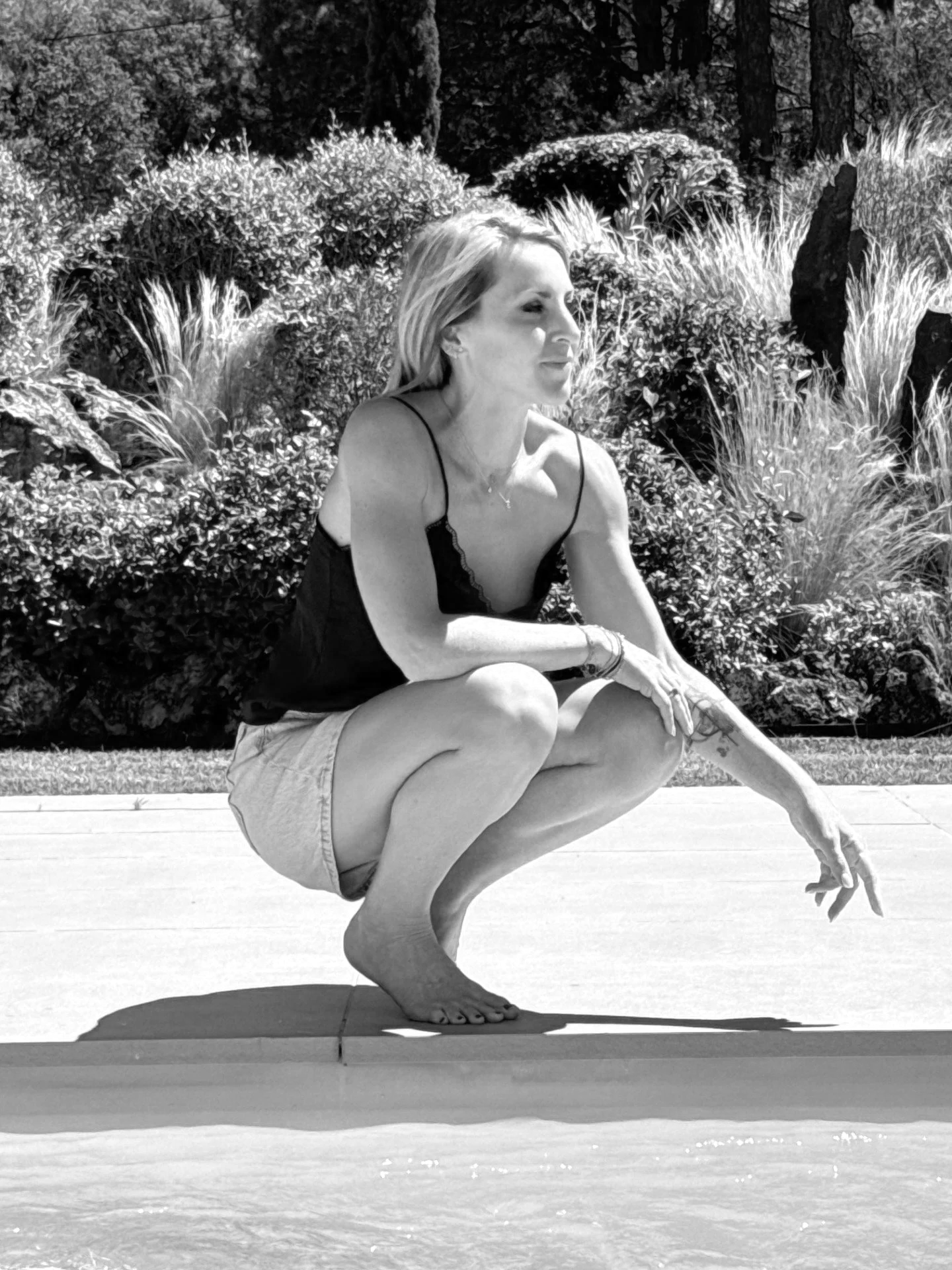 Femme en position de yoga en équilibre sur un pied, tenant l'autre jambe pliée avec une main, sans chaussures, en extérieur face à un mur végétal.