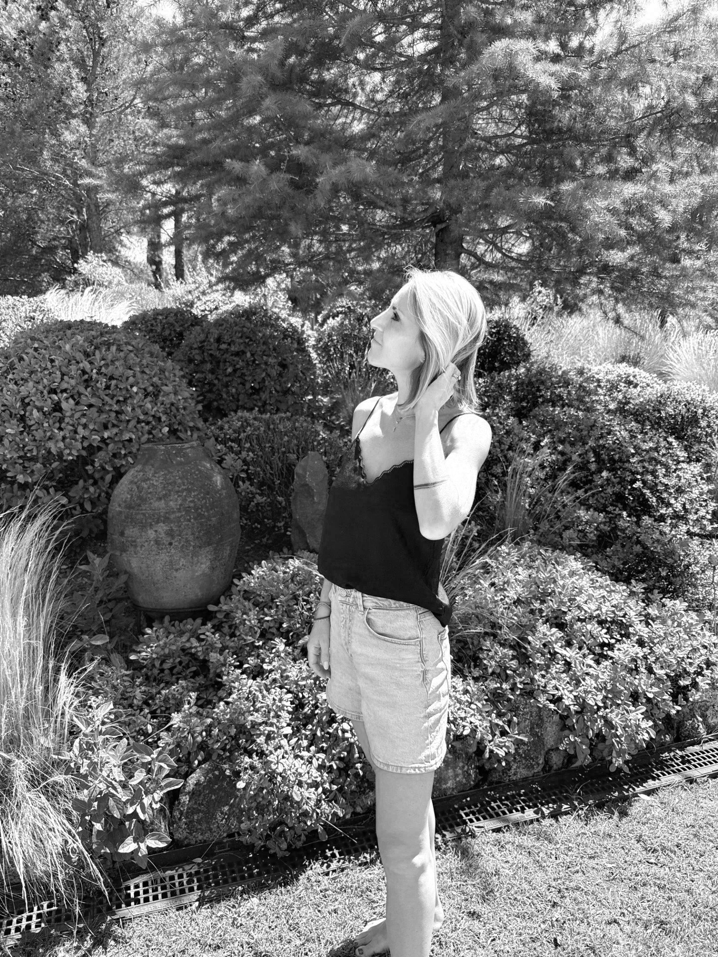Femme debout dans un jardin, entourée d'arbustes et de plantes, en train de se toucher la nuque, en noir et blanc.