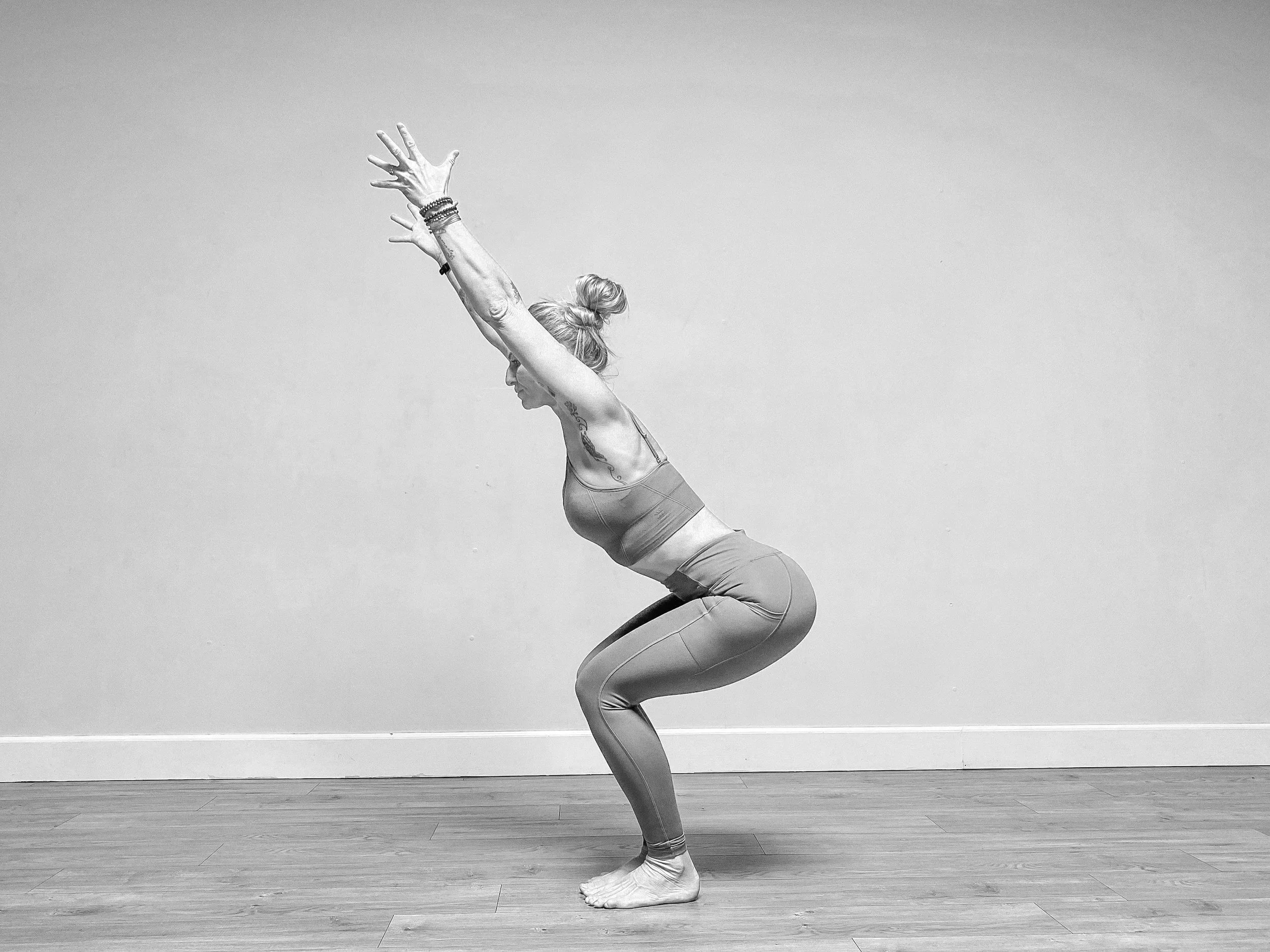 Une femme pratique une posture de yoga ou de méditation dans une pièce avec un sol en bois et un mur gris. Elle est en position accroupie avec les bras levés au-dessus de la tête, les mains ouvertes et les yeux fermés, portant un bandeau ou un chignon en cheveux gris.
