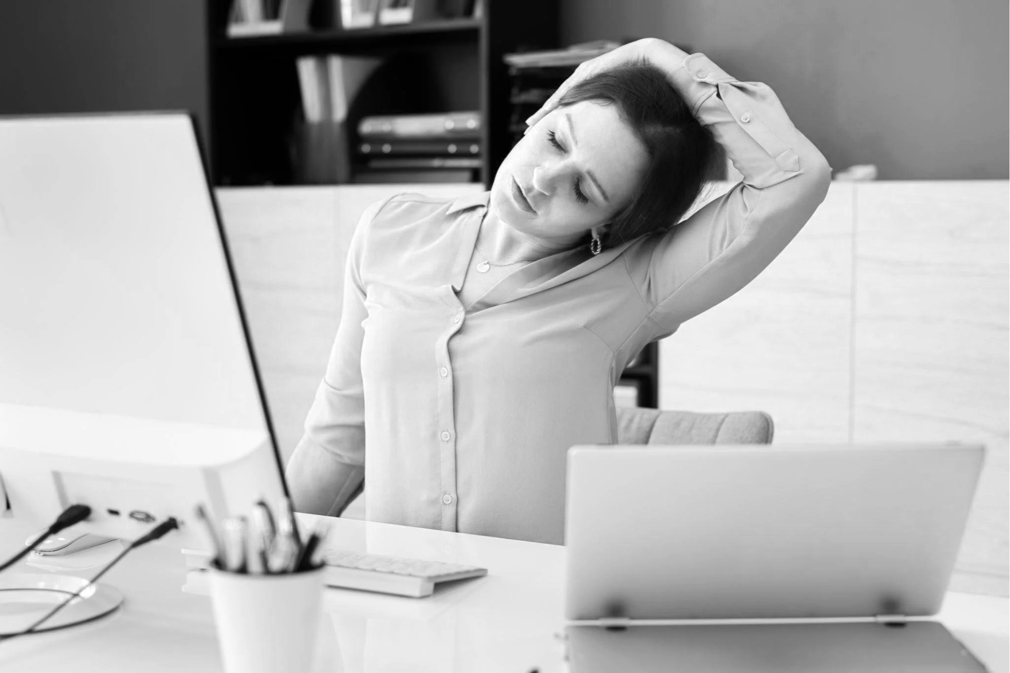 Femme d'âge moyen en chemise claire, assise à un bureau en train de se fatiguer ou de s'étirer, dans un environnement de bureau.