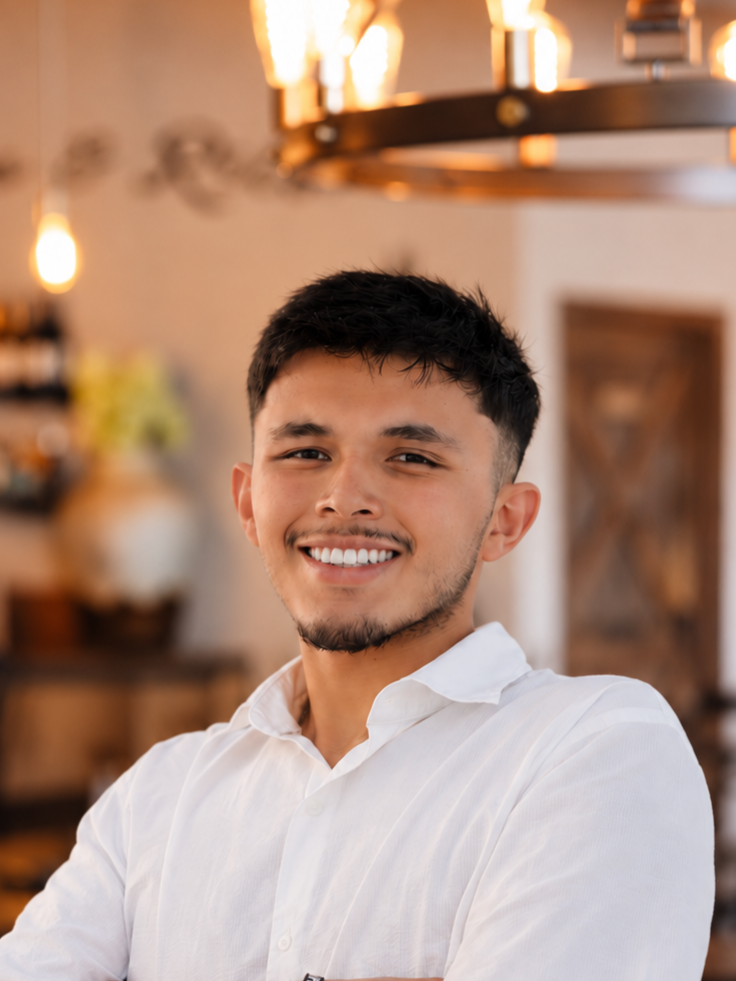 A young man with short dark hair and facial hair, wearing a white collared shirt, smiling in a warmly lit indoor setting with blurred background decor.