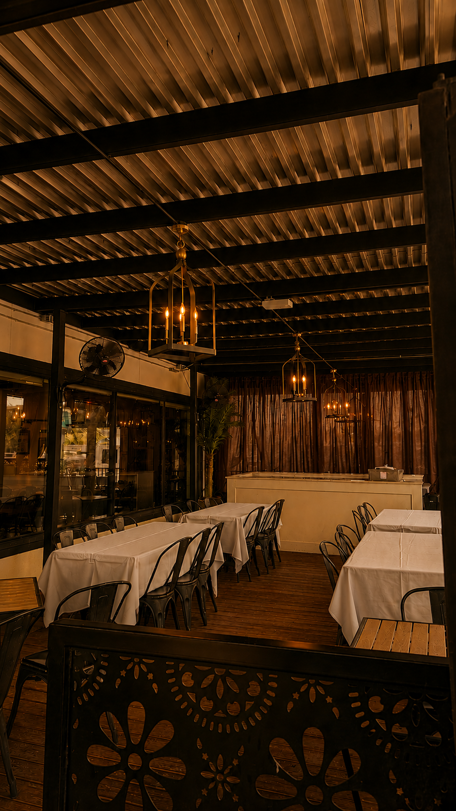 Empty restaurant with white tablecloth-covered tables, black chairs, wooden floor, and warm lighting from chandeliers with candles.