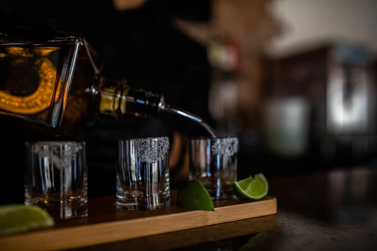 Pouring tequila into shot glasses on a wooden tray with lime wedges in the foreground.