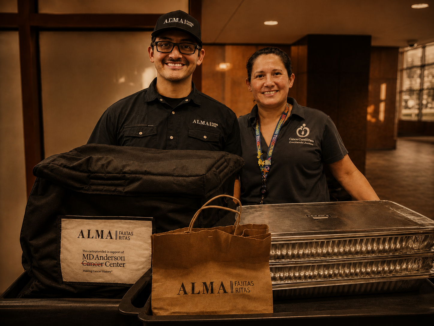 Two smiling people standing behind a cart with food supplies in a lobby. The man on the left wears glasses, a black cap, and a black shirt with "ALMA" embroidered on it. The woman on the right wears a dark shirt with a lanyard. There are bags and a chafing dish on the cart, supporting a cause for cancer care and making cancer history.