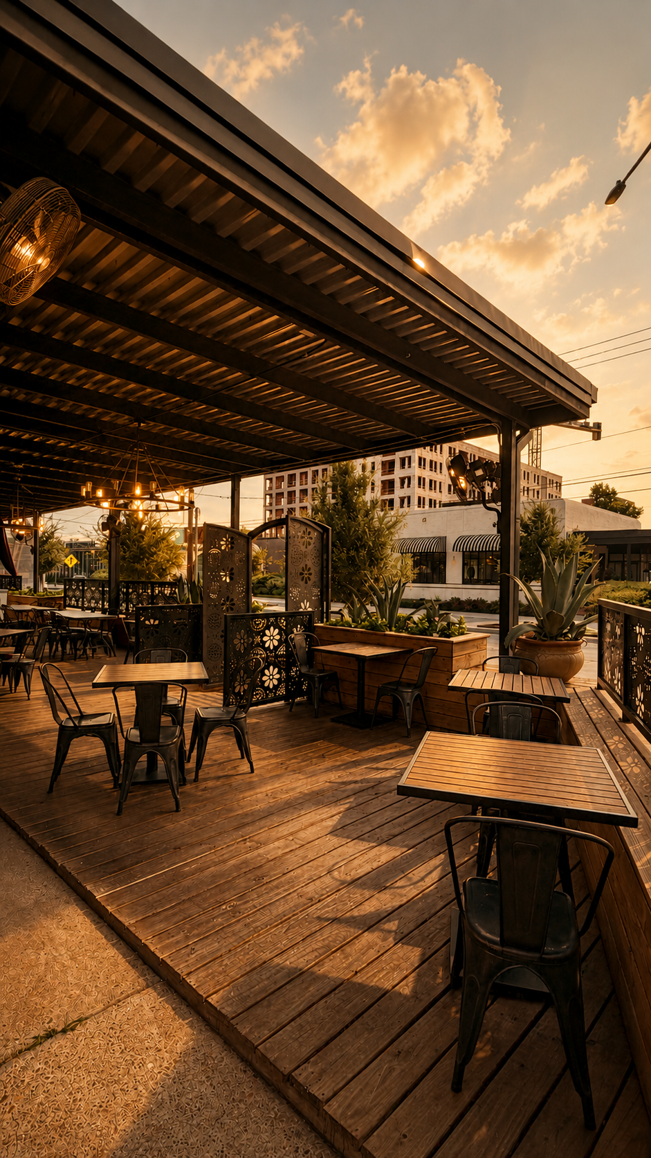 An outdoor dining area with wooden tables and black metal chairs under a roof, with decorative room dividers, potted plants, and warm sunset lighting.