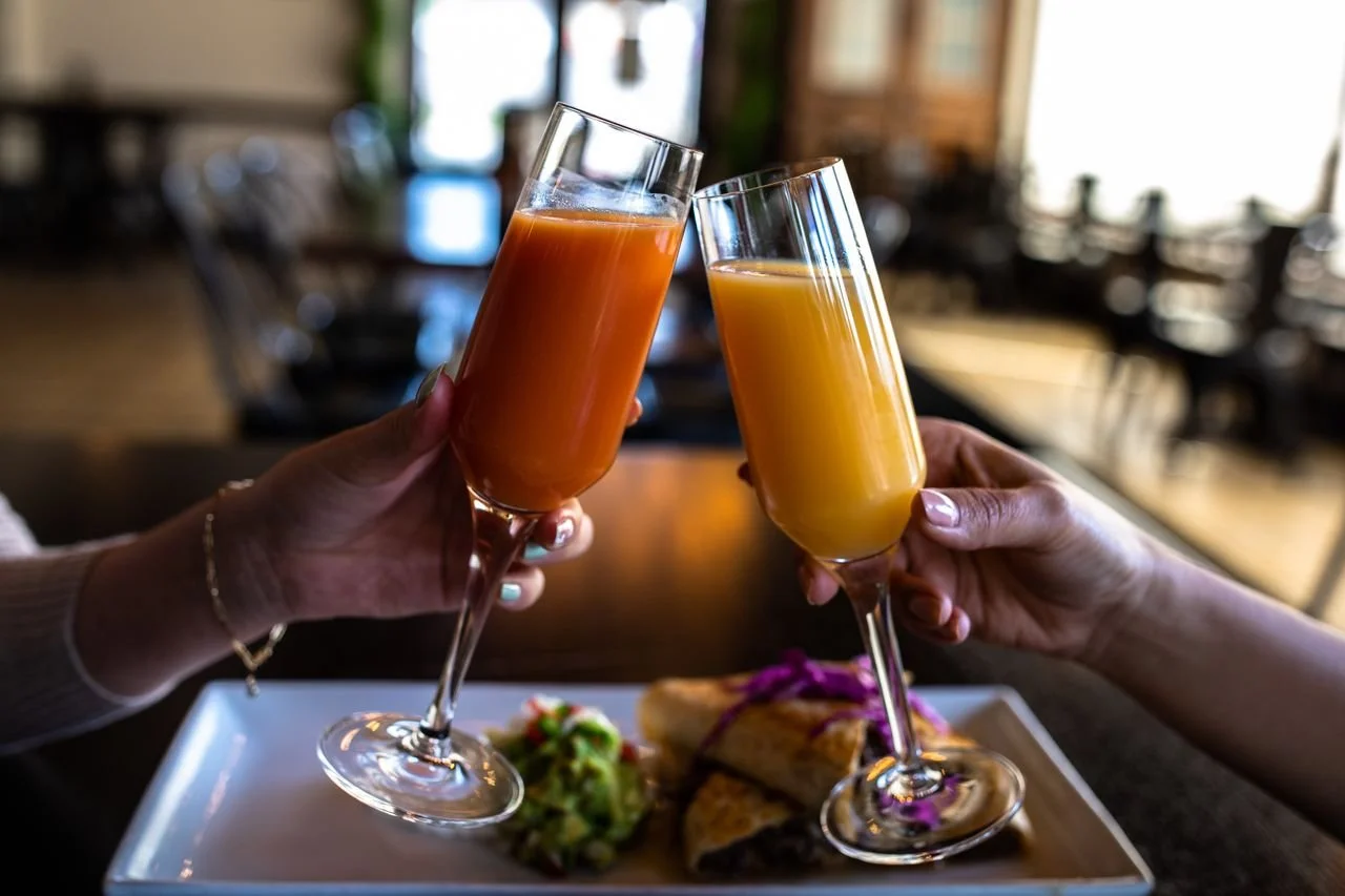Two people clinking glasses of orange juice at a restaurant table with a plate of food in the background.