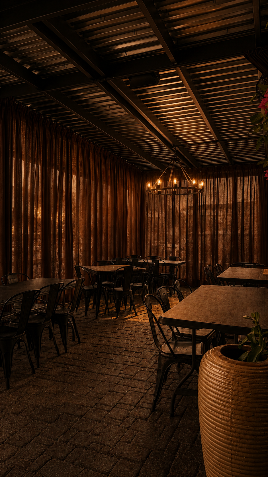Dimly lit outdoor dining area with wooden tables and metal chairs, gold curtains, a large pot planter, and a chandelier with candle-like lights, creating a warm ambiance.