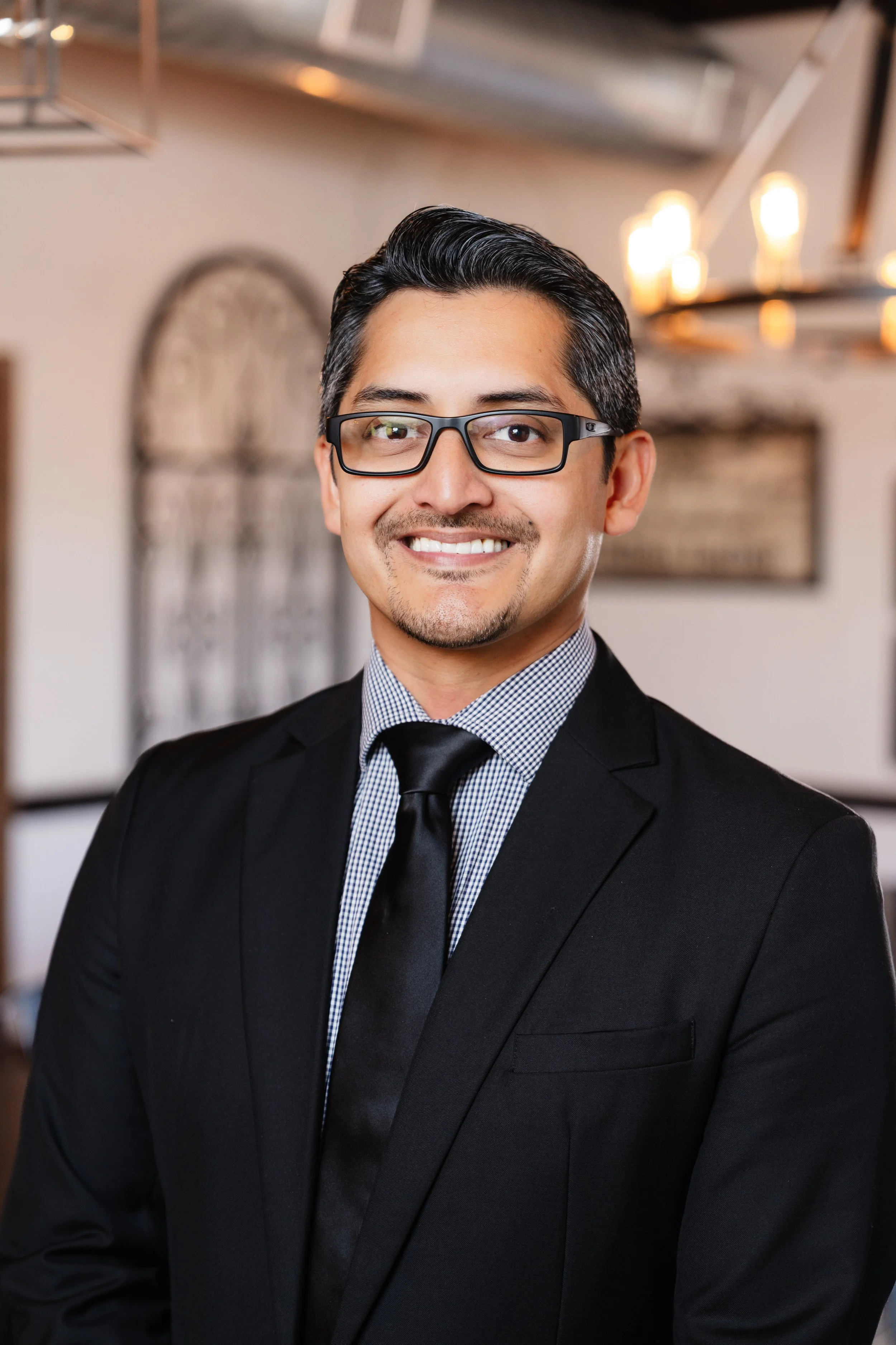 Headshot of a man with glasses in business attire, smiling in an indoor setting with warm lighting and blurred background.