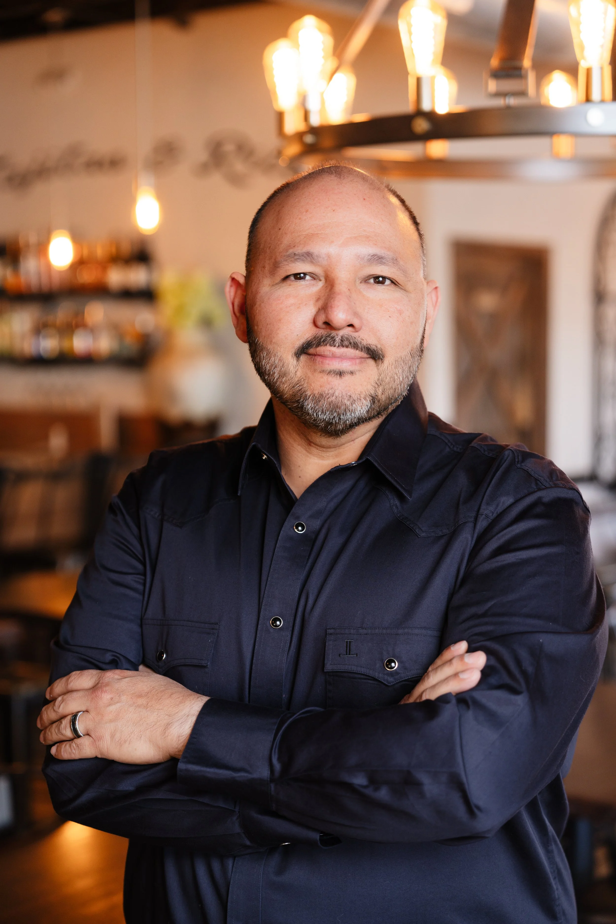 A man with a beard and mustache, wearing a dark navy blue shirt with crossed arms, stands in a warmly lit cafe or restaurant with hanging light fixtures and blurred background furniture.