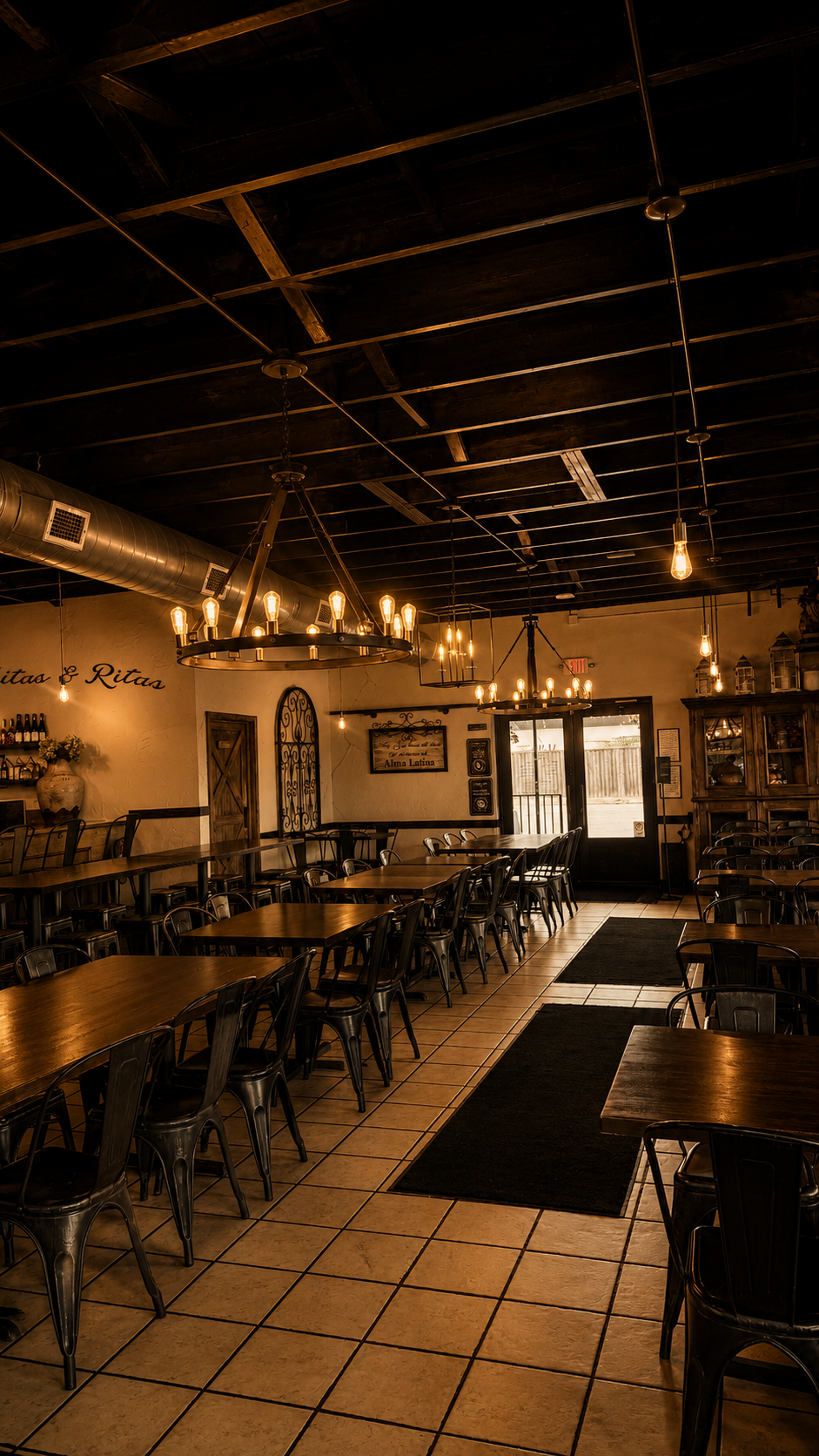 Interior of a cozy restaurant with wooden tables, black metal chairs, warm lighting fixtures, and a tiled floor.