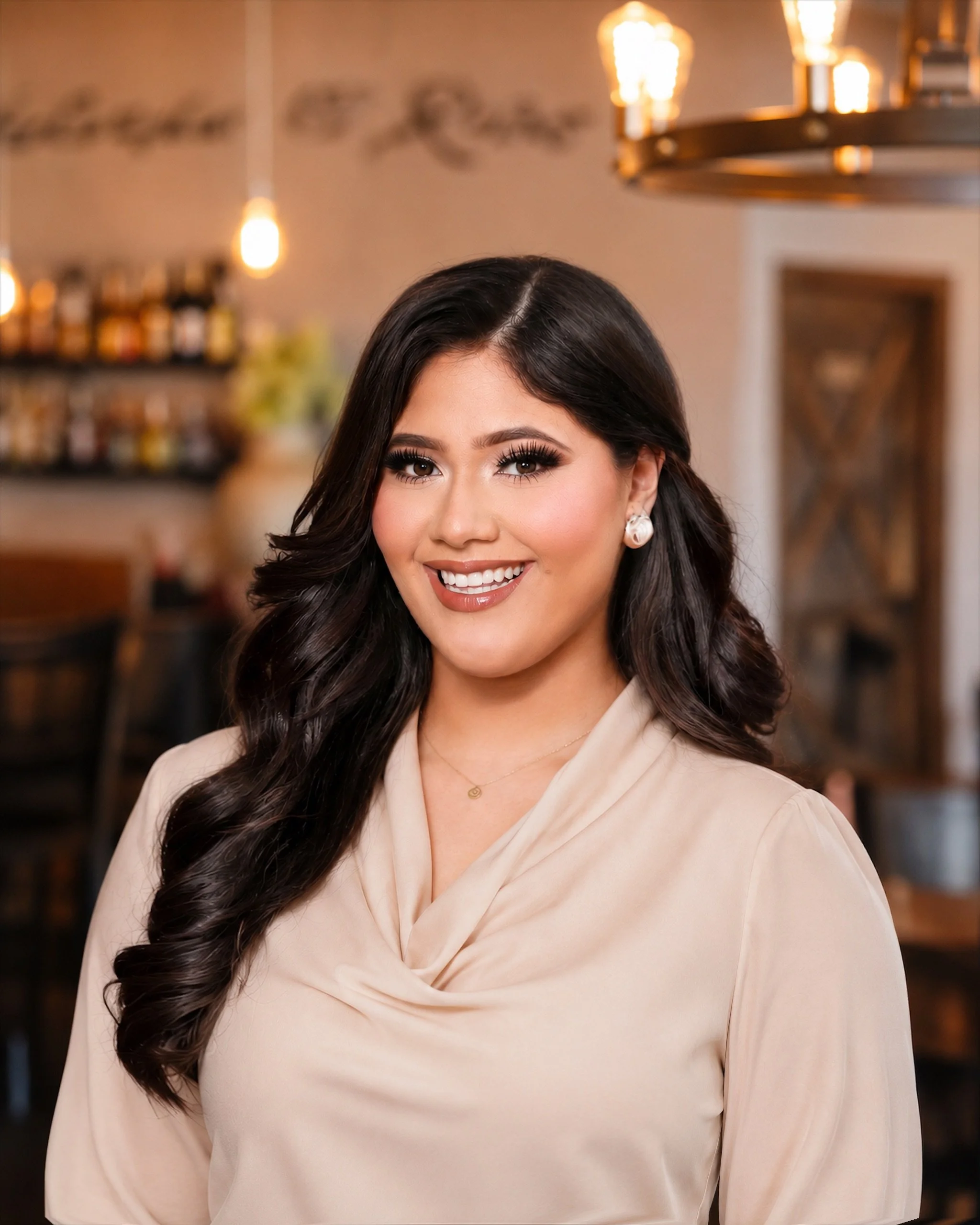Close-up portrait of a woman with long dark hair styled in loose curls, wearing makeup with dark eyeliner and mascara, earrings, and a beige top with a draped neckline, smiling in an indoor setting with warm lighting and blurred background.